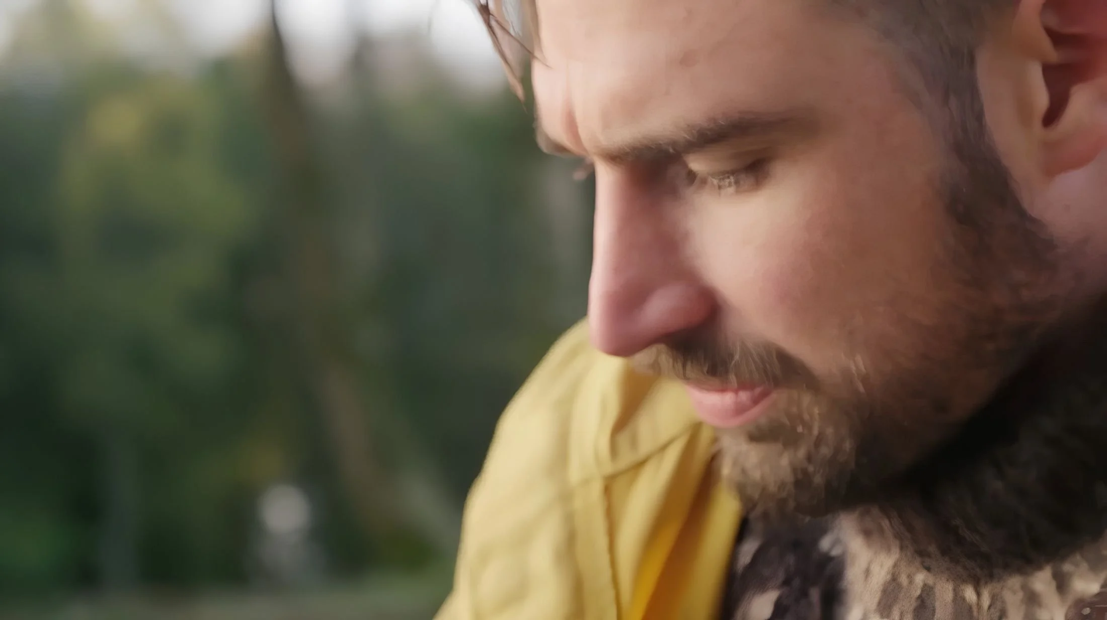Close-up of a man with a beard and mustache, looking downward with a serious expression, outdoors with blurred greenery in the background.