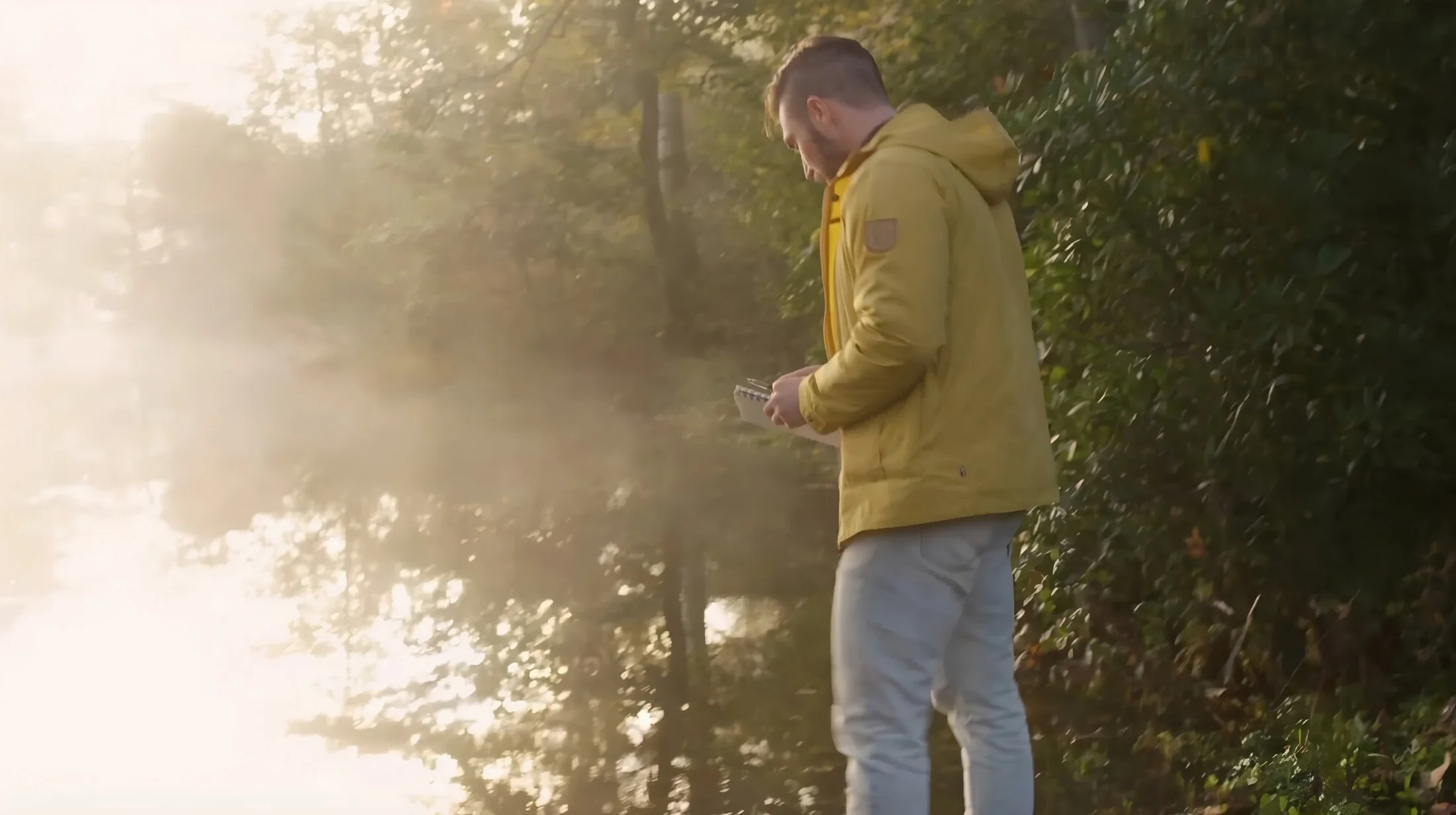 A man with a yellow jacket and light-colored pants stands outdoors amongst trees during sunset, looking at his phone.