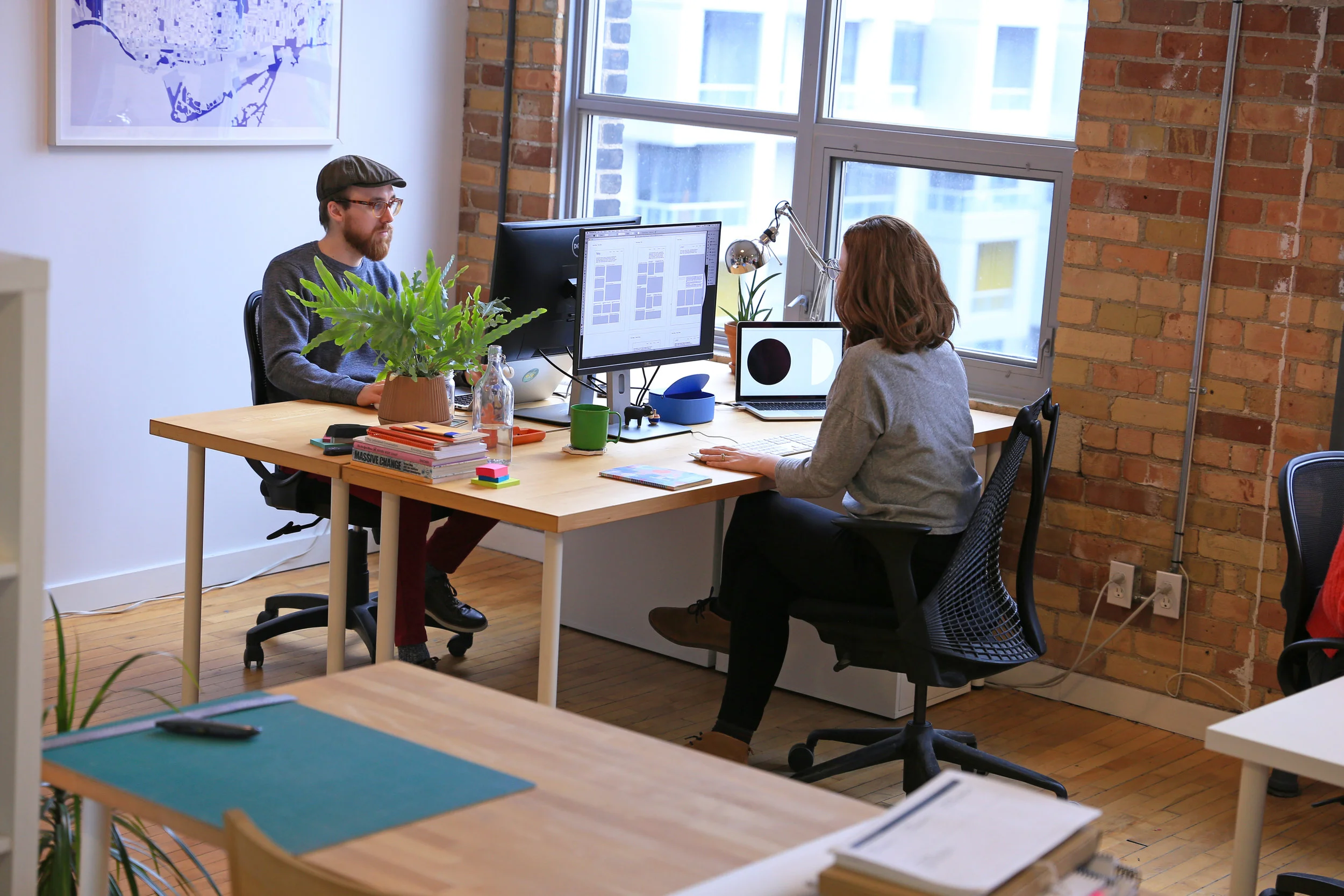 Jay and colleague Kaila work on their computers in a studio with brick walls, hardwood floors, and plants around their desks.