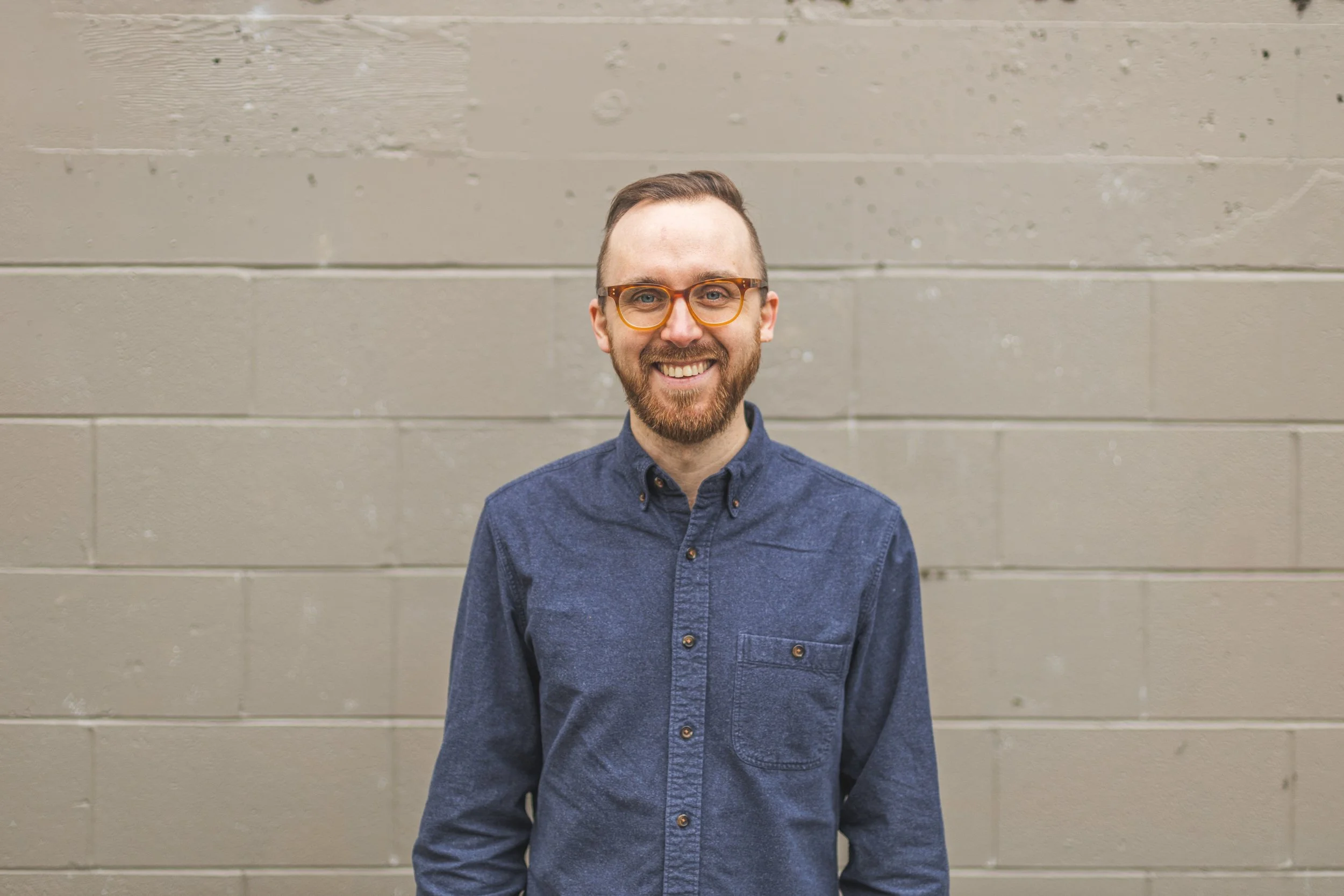 Jay stands in front of a grey wall, smiling toward the camera. He is wearing a navy collared shirt and glasses.