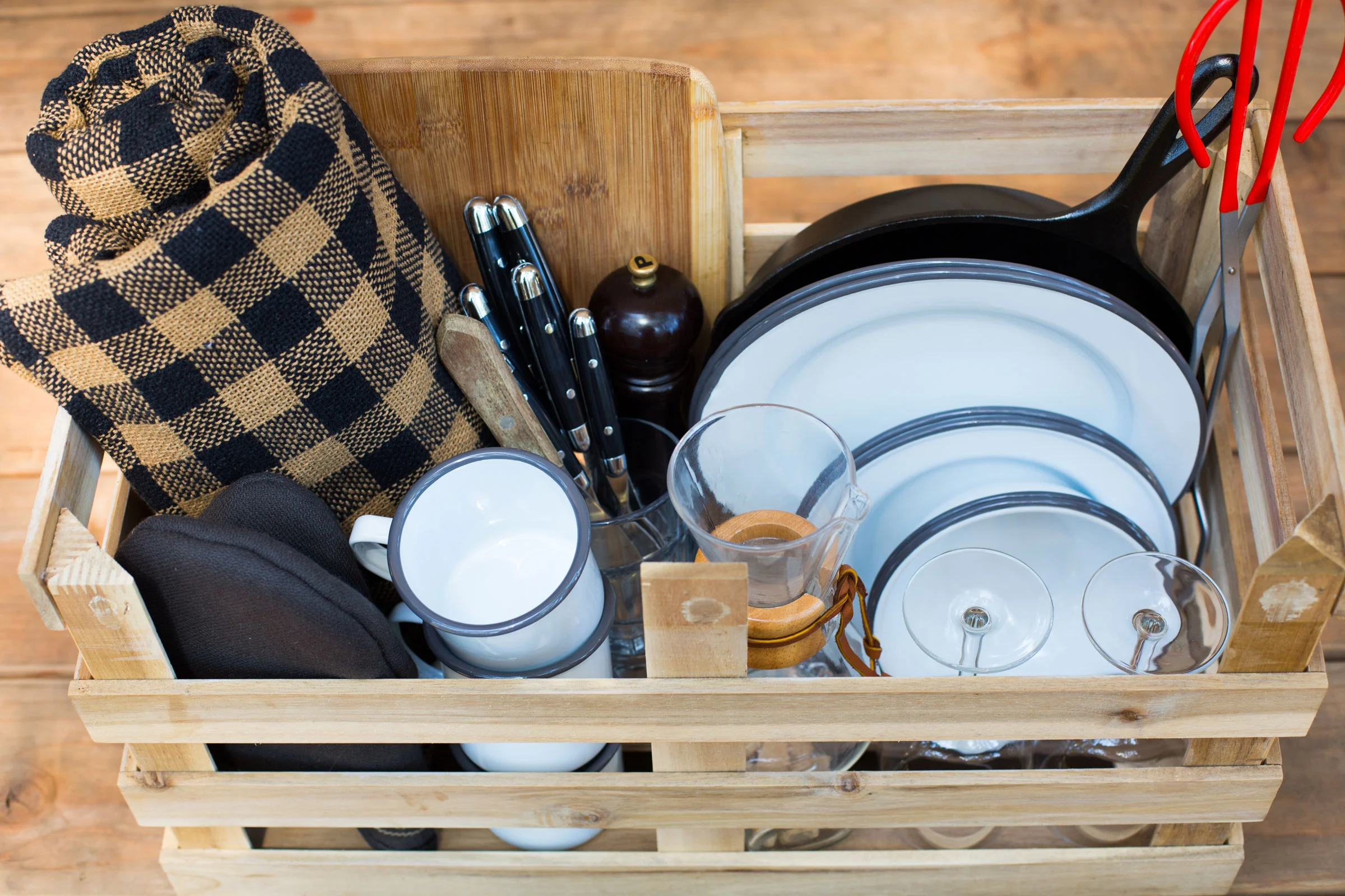 a crate with picnic essentials — metal camping plates and mugs, camping utensils, wine glasses, blanket, pan and more