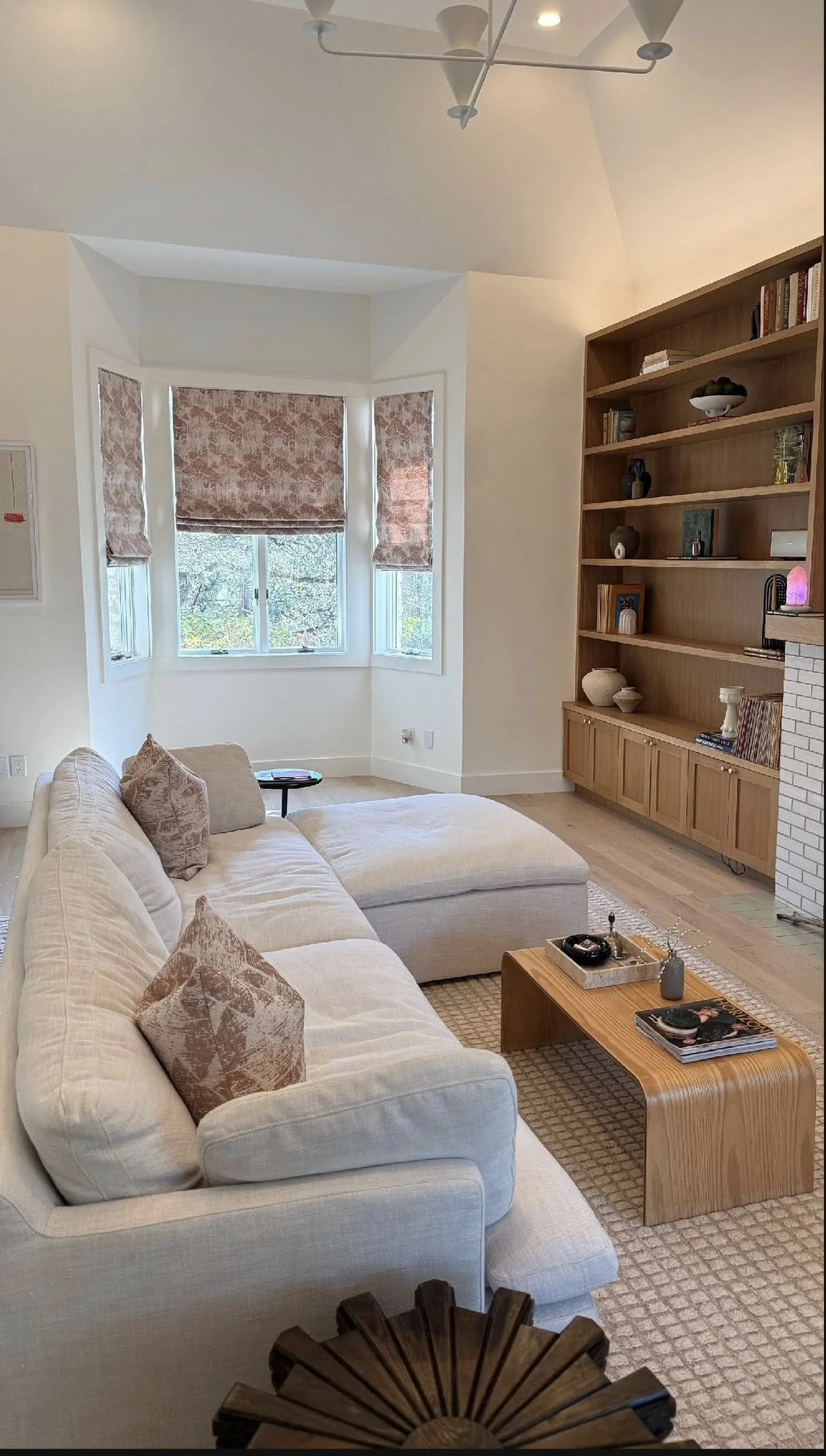 Living room with a light-colored sofa with patterned pillows, a wooden coffee table with magazines and decorative items, and a tall wooden bookshelf filled with various decorative objects and books. There is a large bay window with pink-brown patterned curtains that are partly open, letting in natural light. The floor is light-colored wood, and there is a textured beige rug. The ceiling has modern light fixtures.