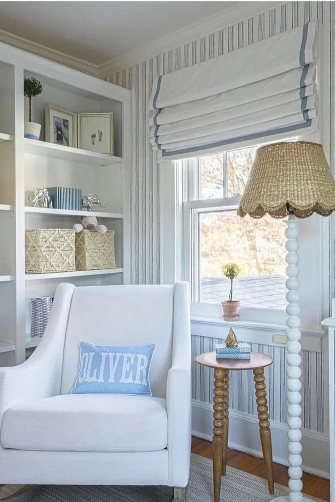 A cozy living room corner with a white armchair that has a blue pillow with the name 'Oliver', a small wooden side table with a blue gift box and a decorative brass object, a tall lamp with a woven shade, a window with white blinds, a built-in shelf with decorative boxes and pictures, and a window sill with a small potted plant.