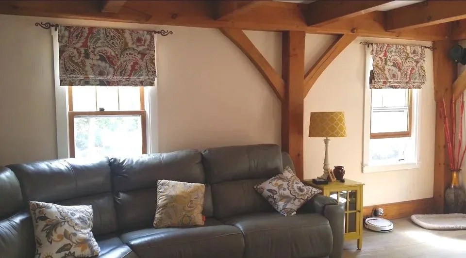 Living room with a gray leather sofa, patterned throw pillows, windows with patterned curtains, a yellow side table with a lamp, a small robot vacuum on the floor, a pet bed, and decorative sticks in a vase.