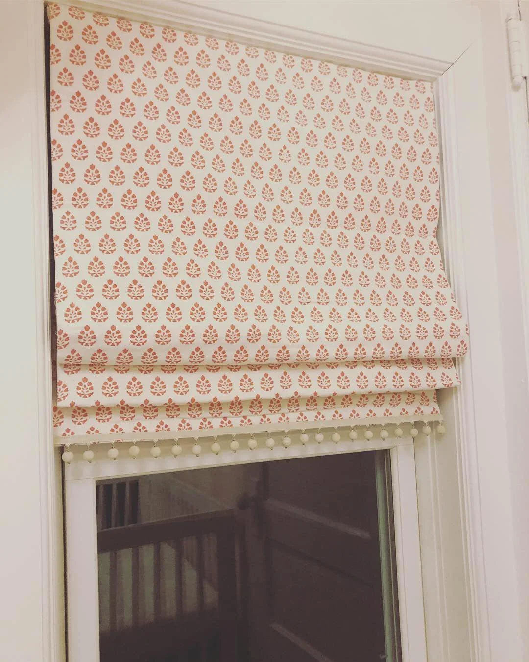 Decorative window blind with a pink and white pattern featuring small floral or leaf motifs, mounted inside a white window frame with molding.