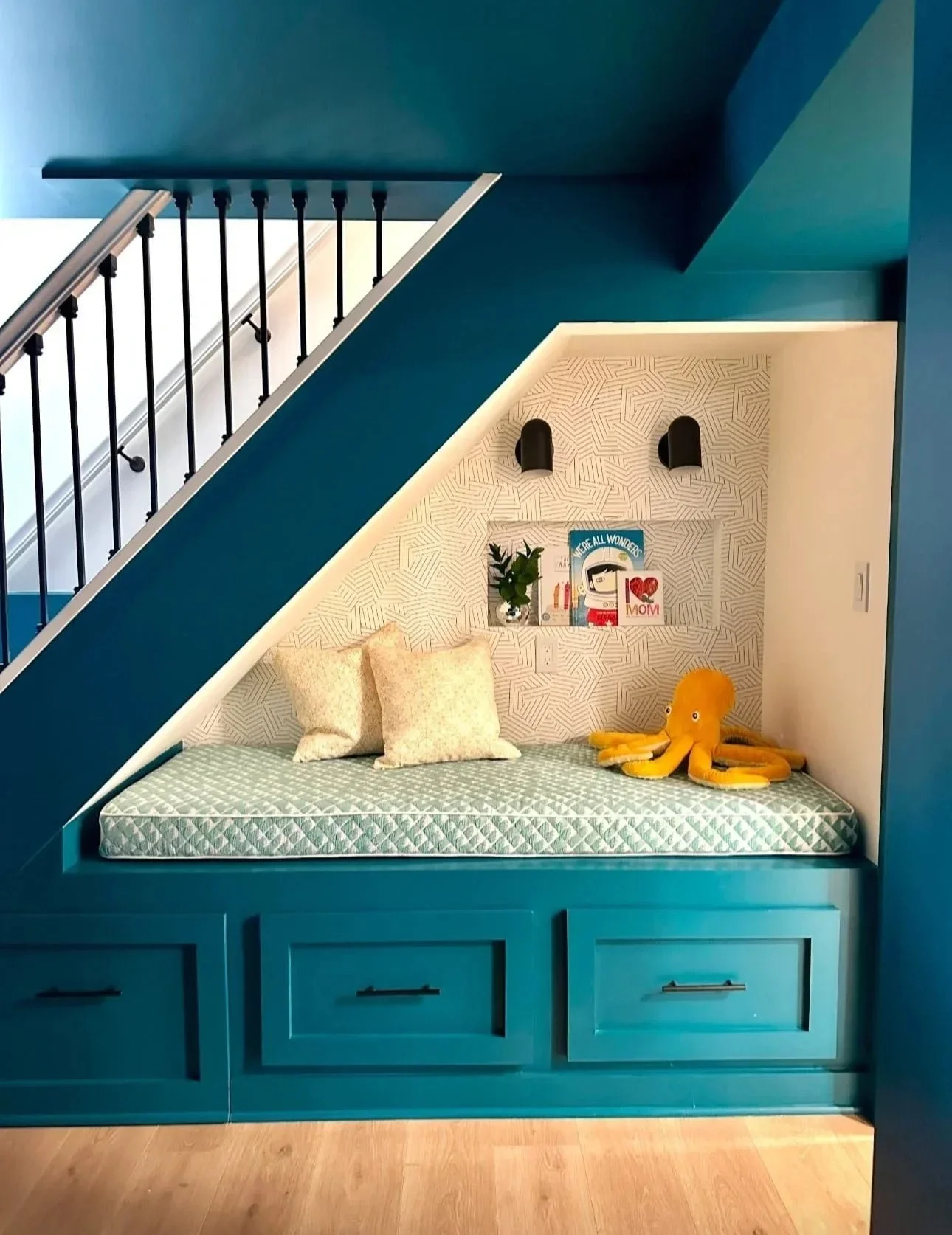 Built-in blue daybed with a patterned mattress, pillows, and plush octopus toy, set under a staircase with black railings, against patterned wallpaper and decorative wall elements.