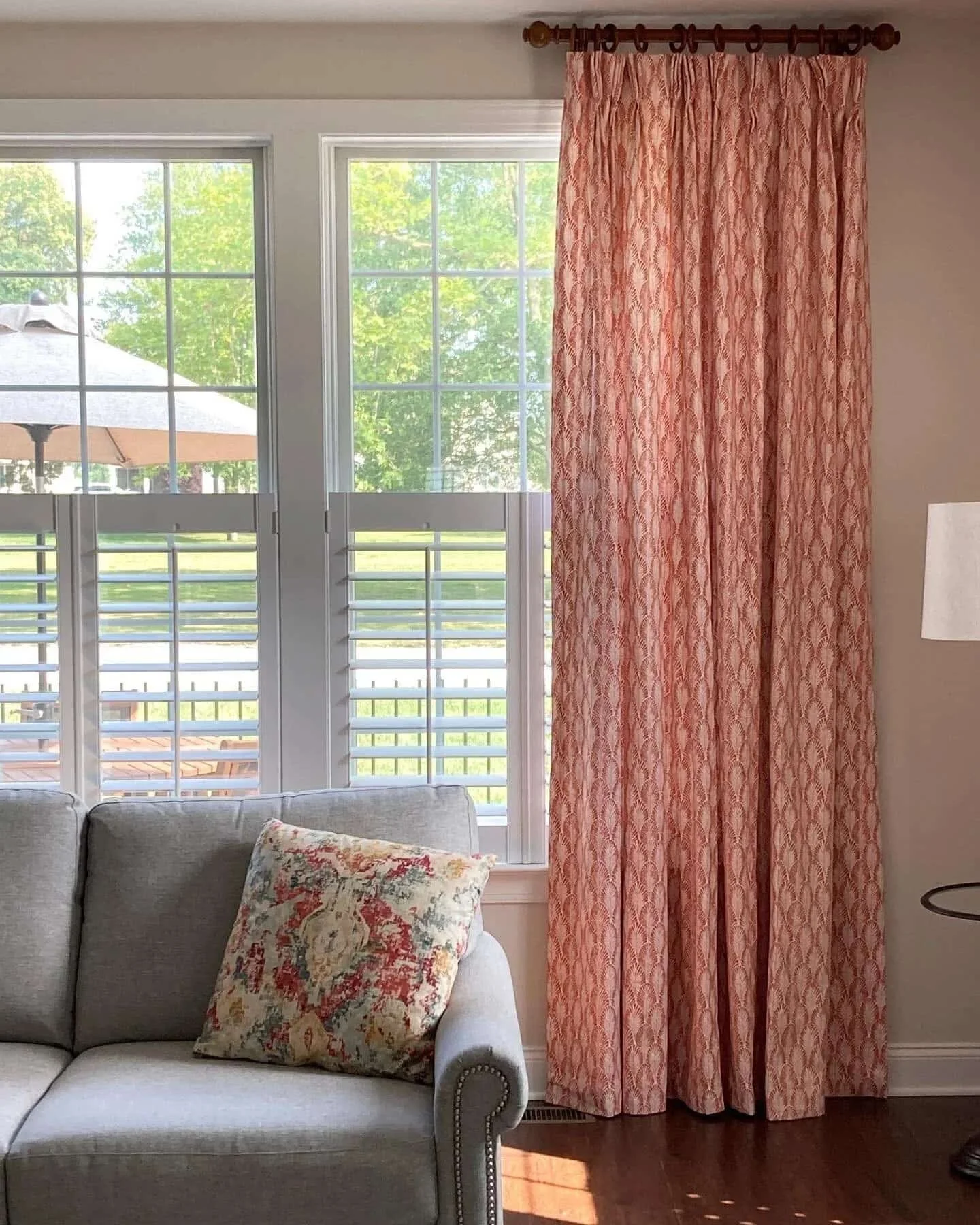 Living room with two large windows with white shutters, pink patterned curtains, a grey sofa with a floral cushion, wooden floor, and a lampshade on the right.