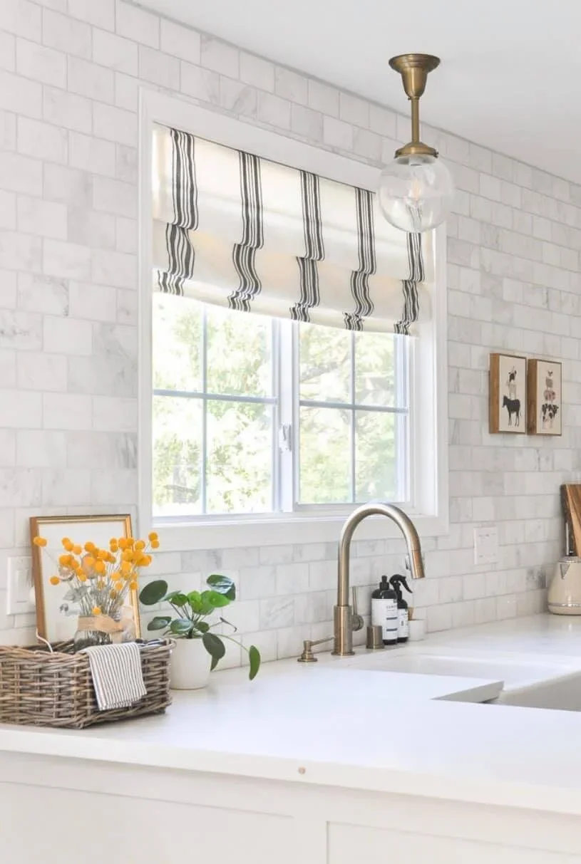 Bright kitchen with white brick subway tile backsplash, a window with a black and white striped roman shade, a brass pendant light, a wicker basket with flowers, a green potted plant, and some soap dispensers on the white countertop.