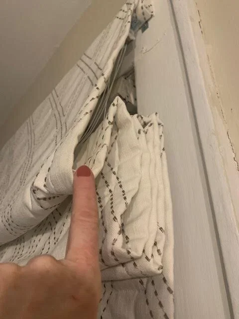 Close-up of a finger pointing at a small gap between a white curtain and a white window frame.