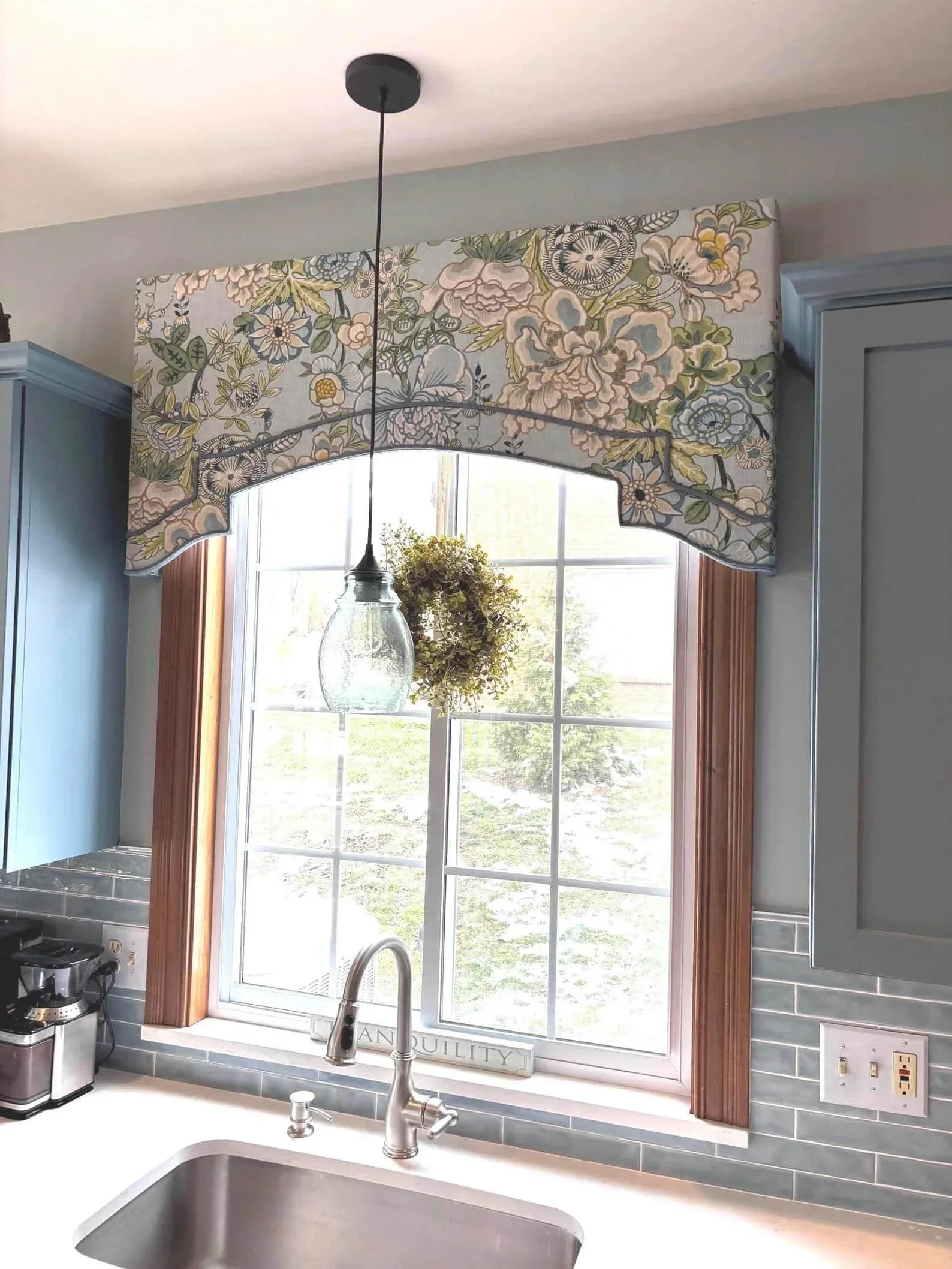 Close-up of a kitchen window with floral patterned valance, a hanging glass pendant light, a flower arrangement, a sign that reads 'tranquility,' a blue kitchen cabinet, a gray tiled backsplash, and a kitchen sink with a silver faucet.