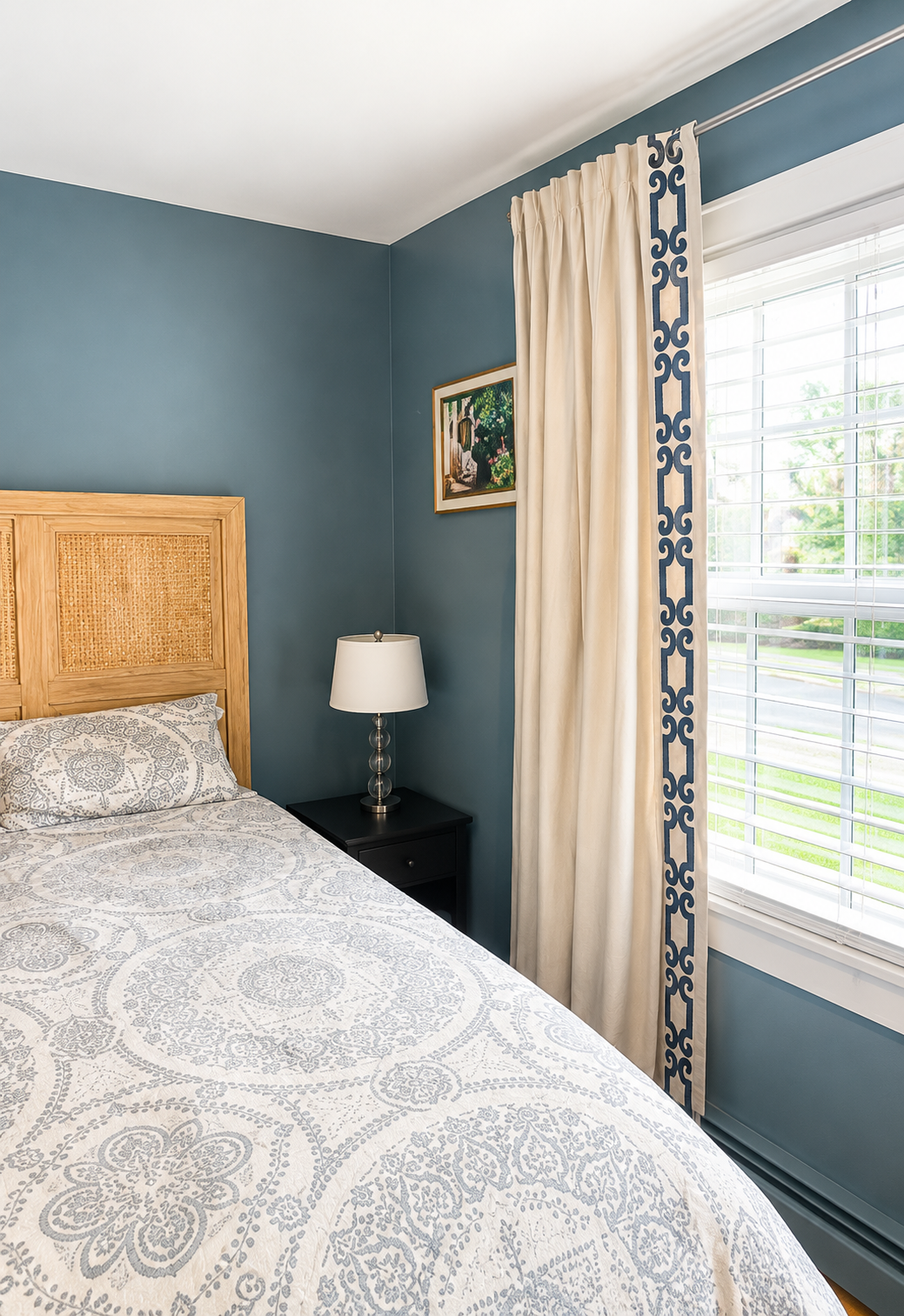 A bedroom with blue walls, a bed with a patterned white and gray comforter, a wooden headboard, a black nightstand, a white table lamp, a window with white blinds, and cream-colored curtains with a blue border. A small framed picture hangs on the wall.
