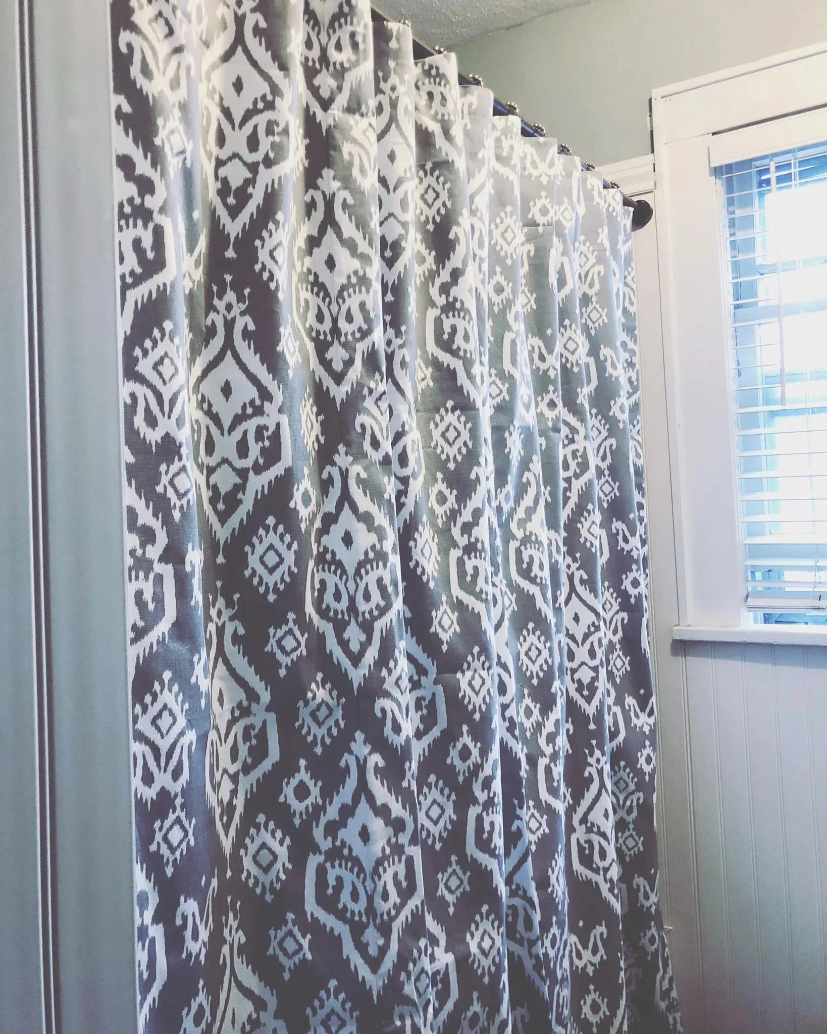 A window with white blinds, and a decorative curtain with a white and gray diamond pattern.