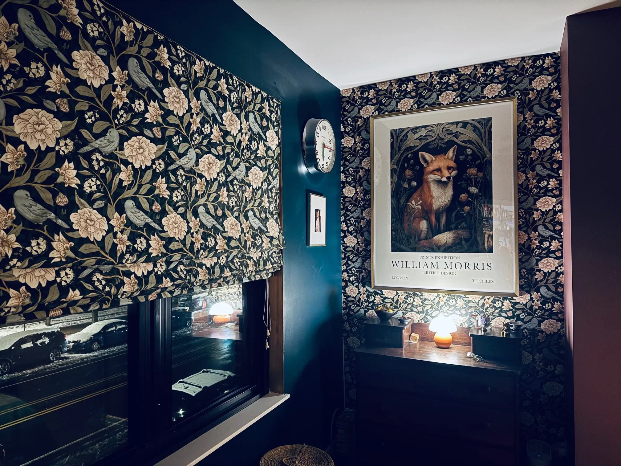 Interior view of a room with floral and bird-patterned wallpaper on the walls. A large framed picture of a fox accompanies a poster about William Morris, a British textile designer, on the wall. There are lamps on a table below the poster, and part of a window showing parked cars and snow outside.
