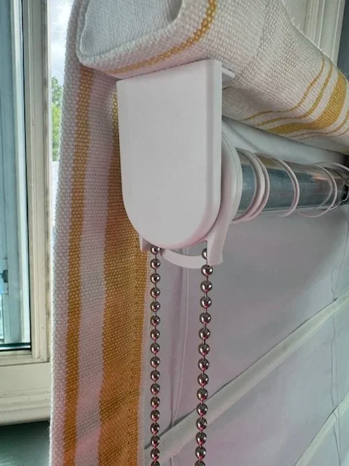 A window with a white curtain partially pulled back, a white roller blind partially rolled up, and a beaded chain pull hanging from the roller blind mechanism.