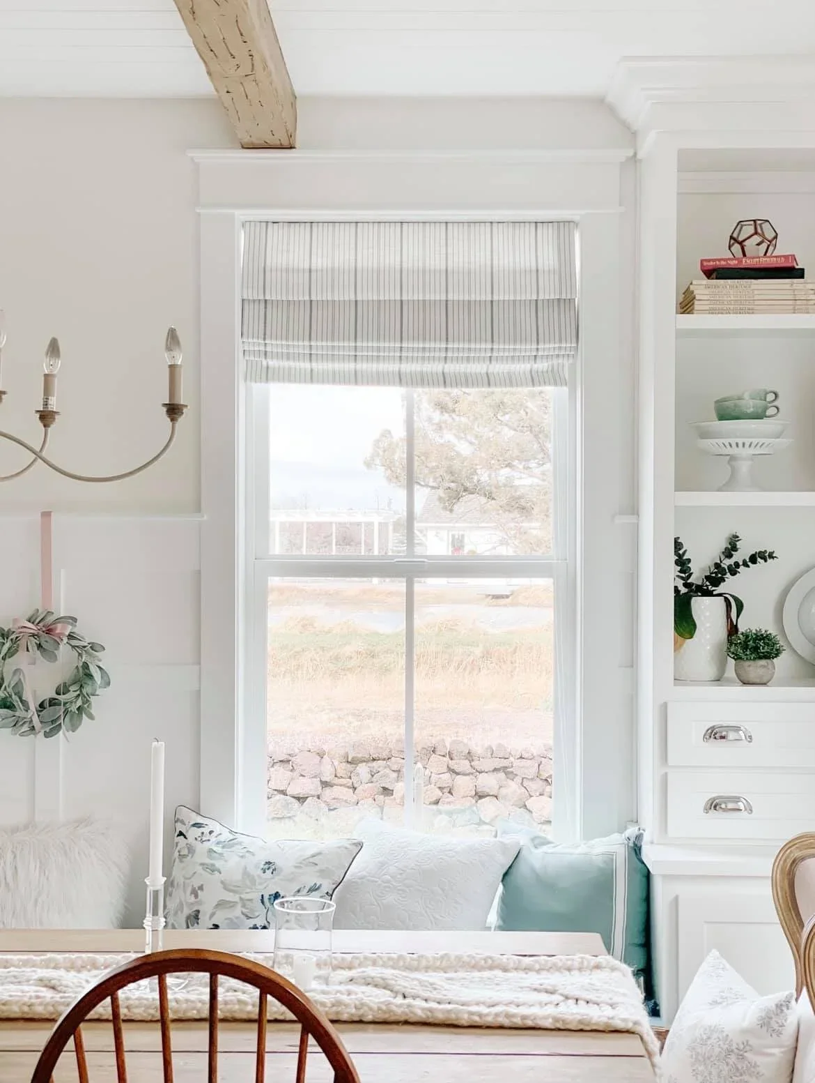 A bright living room with a window dressed in a white and grey striped valance, overlooking a yard with rocks and trees. White built-in shelves contain books, a green bowl, a plant, and decorative items. A chandelier with candle-shaped bulbs hangs on the left.