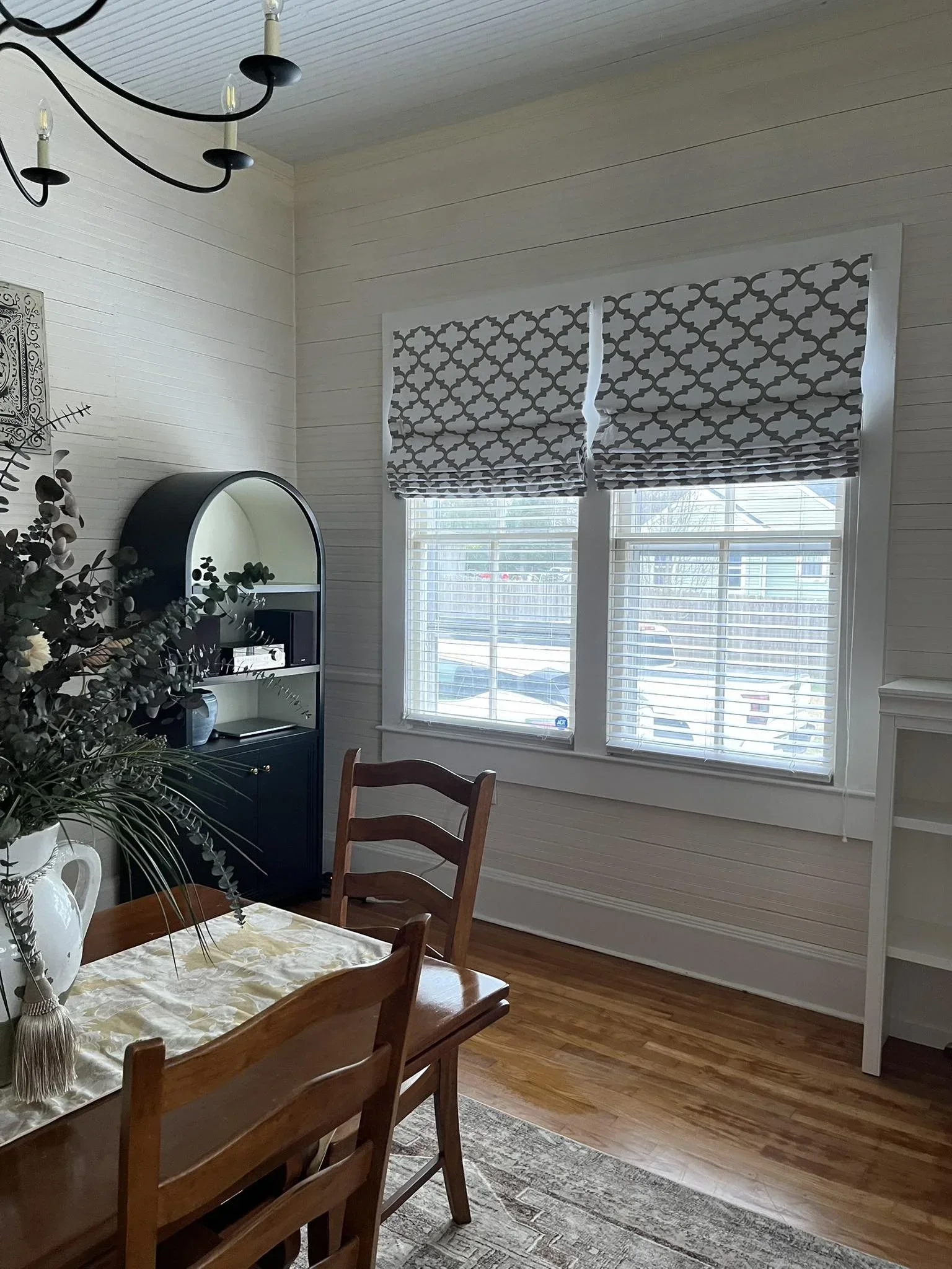A dining room with a wooden table and chairs, a black and white patterned window treatment, beige walls, hardwood floors, a black chandelier, a white bookshelf, and decorative plants.