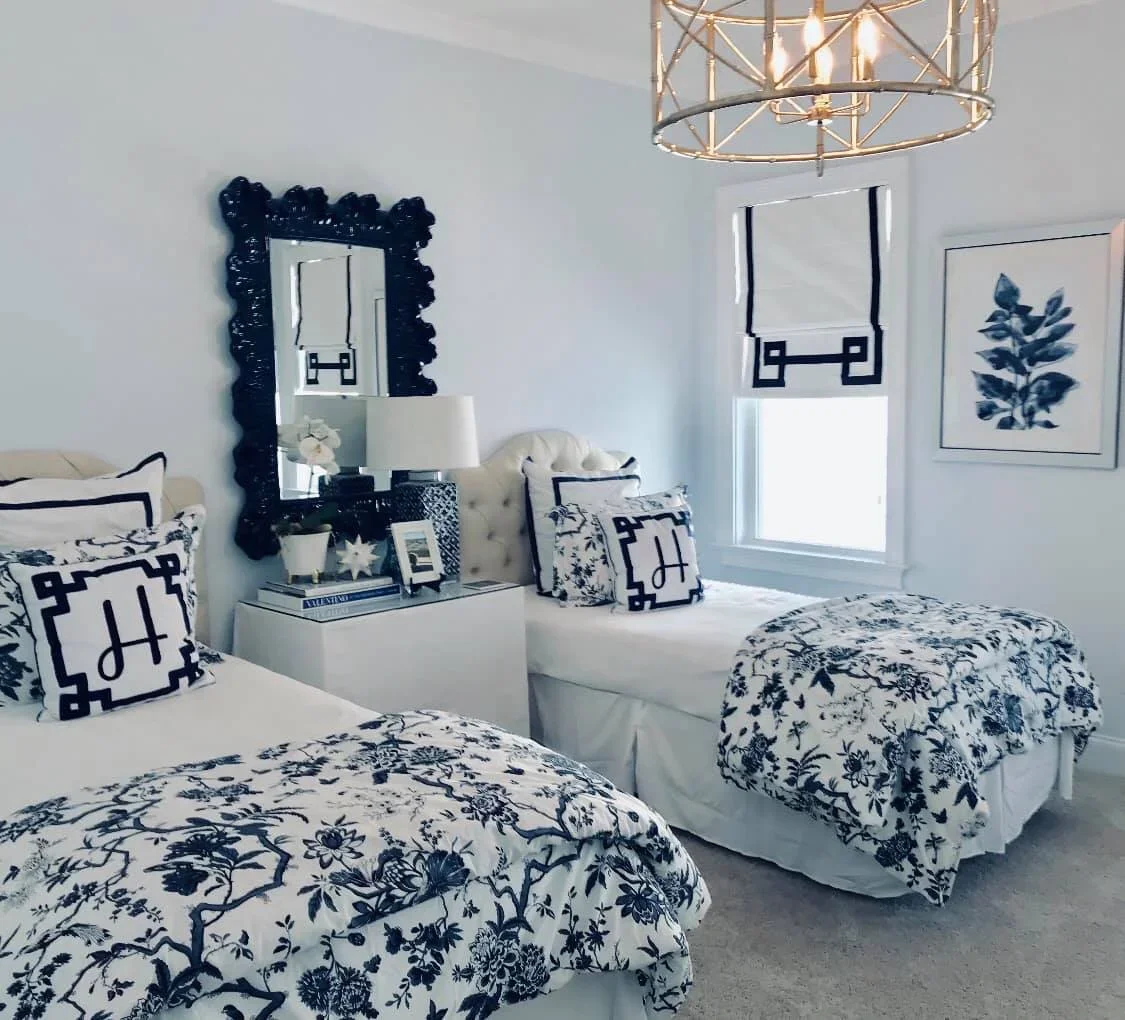 A bedroom with white walls, two beds with black and white floral bedding, a white nightstand with decorative items and books, a cream tufted chair with black and white patterned pillows, a black-framed mirror, a window with black and white curtains, a framed black and white botanical artwork, and a gold chandelier overhead.