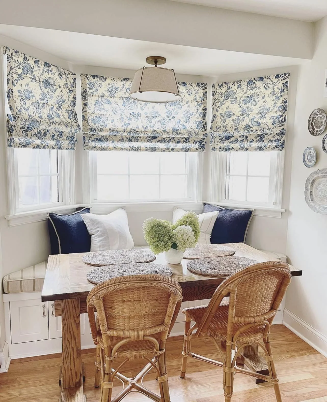 A cozy dining nook featuring a wooden table with woven placemats, surrounded by rattan chairs, and a built-in bench with pillows. Three large windows with floral Roman shades light the space, and a flower arrangement decorates the table. A ceiling light hangs above.