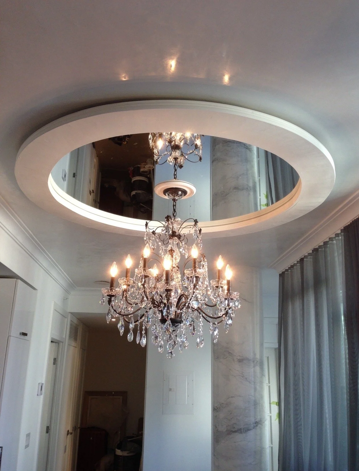 A chandelier hangs from the ceiling in a room with elaborate ceiling design and curtains on the window.