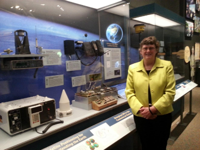 Caption: Dr. Alison Brown, President and CEO of NAVSYS Corporation, standing next to the first GPS enabled cell phone which was developed at NAVSYS and is now on display at the Smithsonian’s Navigation and Time exhibition.