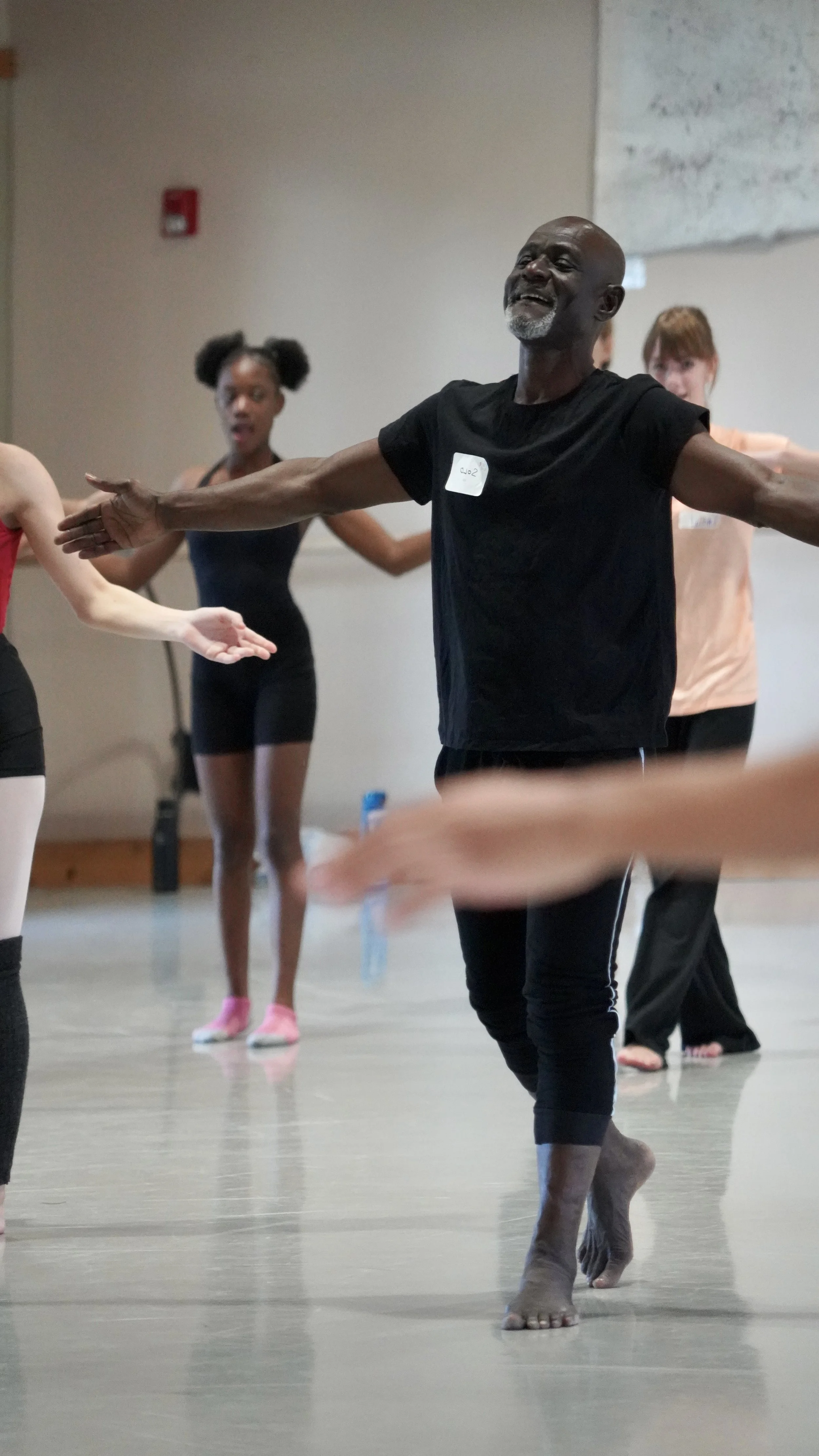 Inside a dance studio with beige walls and a gray floor, a bald, black male teacher with a gray goatee opens his arms wide, chest proud, as he demonstrates a Burkina Faso Dance skill for students, in the background.