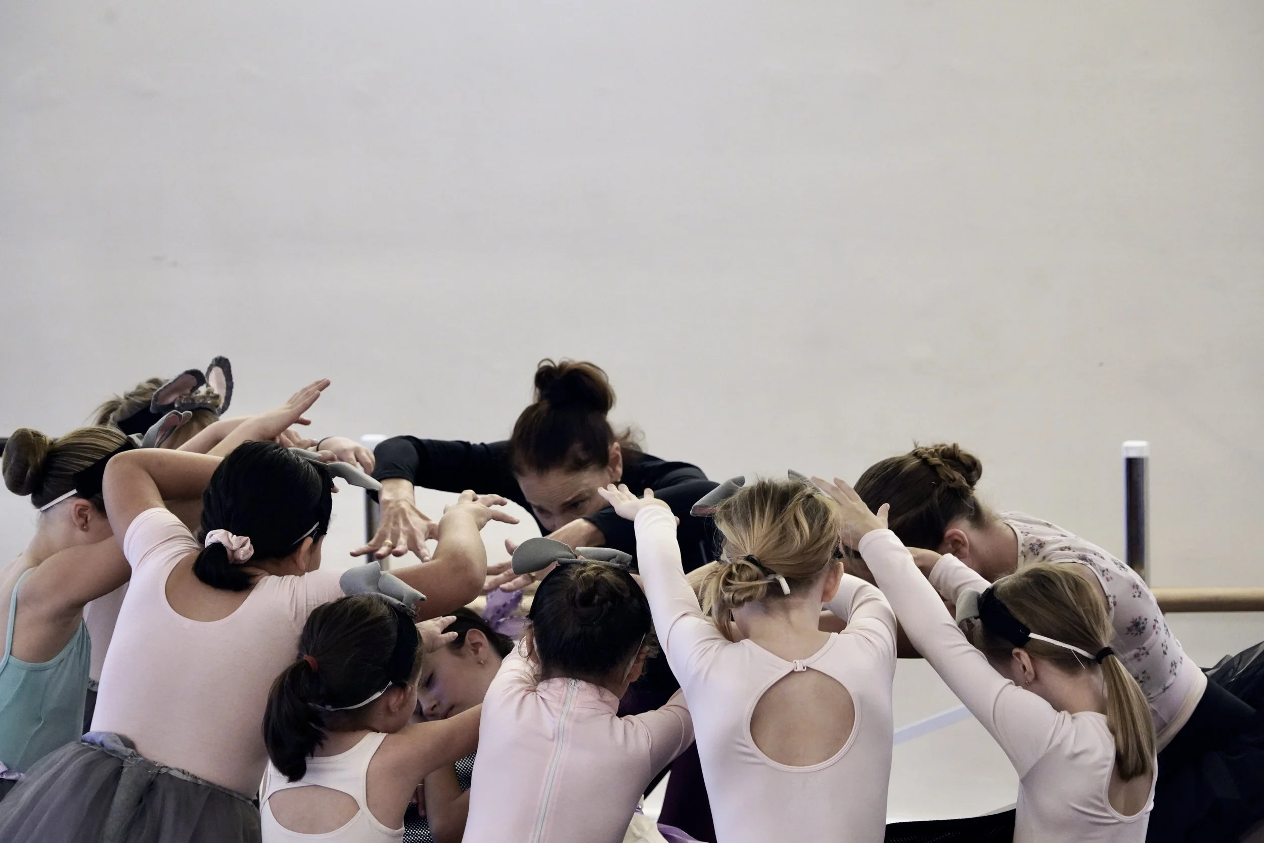 a group of ballet students in pink leotards huddle around their teacher, who is engaged and demonstrating the gesture desired in the choreographic moment