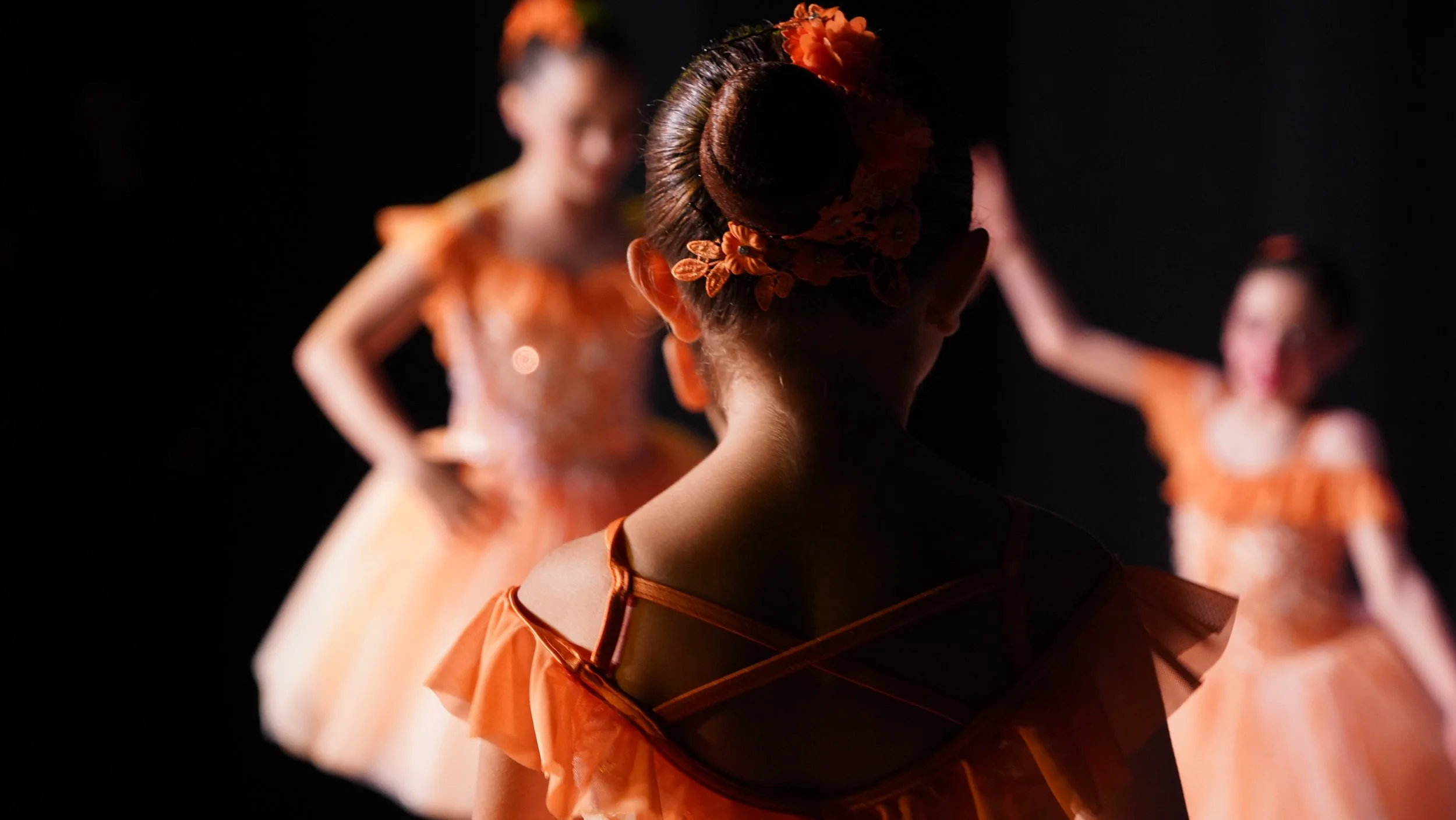 the foreground of the images is the back head and shoulders of a young dance student, adorned in orange performance costume, and with flowers around her hair bun. The background has other youth dancers in the same costume, in various poses.