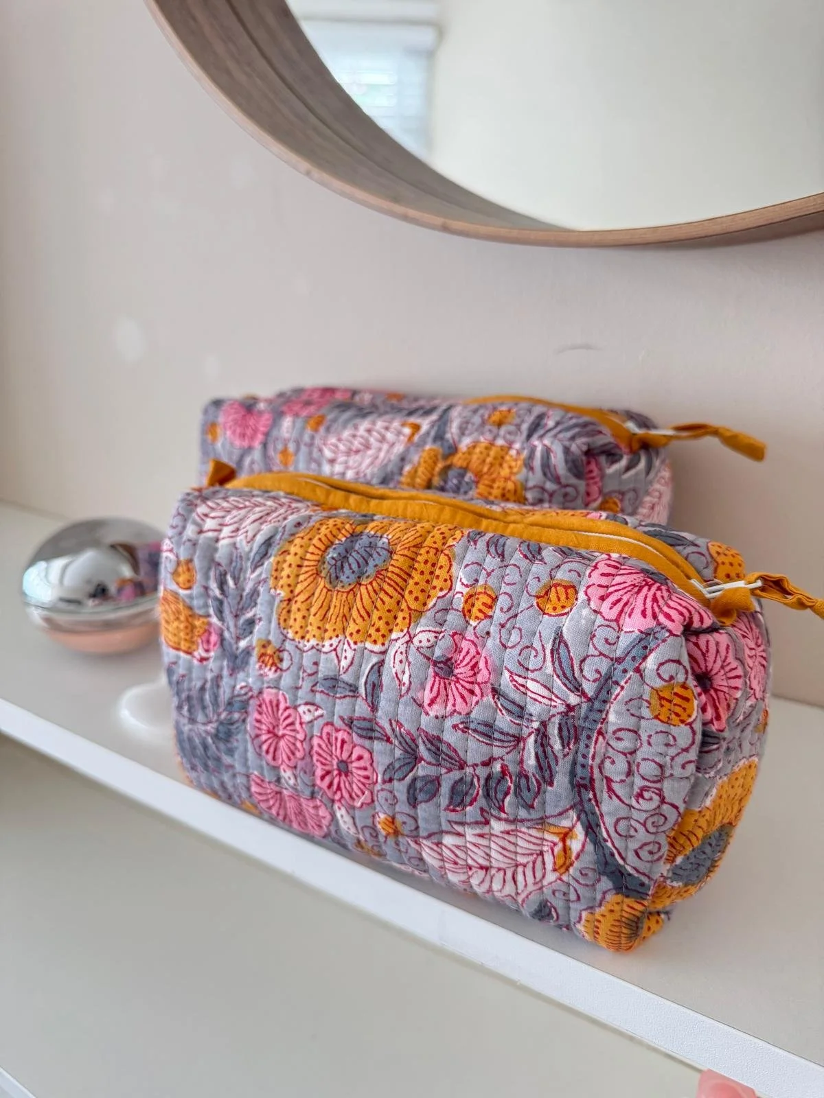 Small and medium size quilted cosmetic bag hand block printed with a floral design, resting on a white shelf under a round mirror