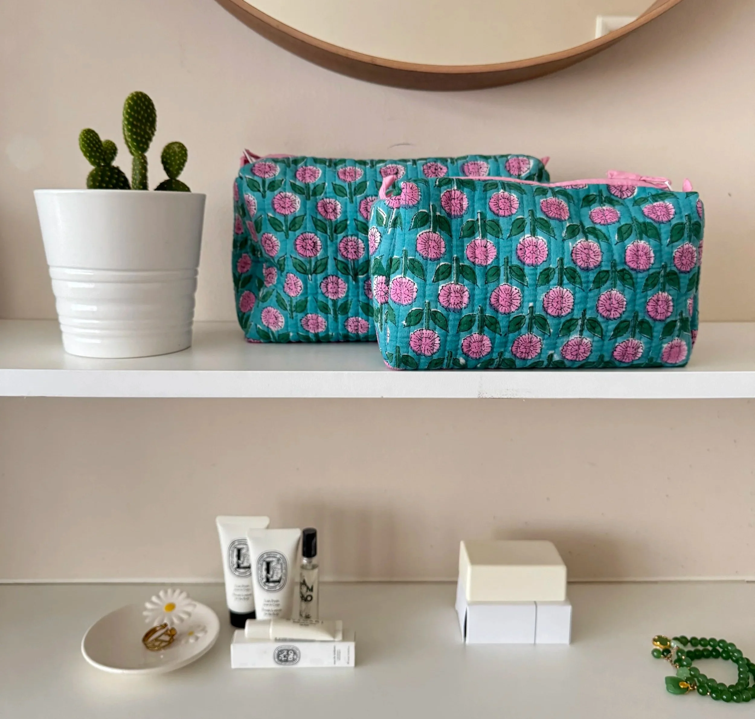 Two block print quilted cotton wash bags resting on a white shelf under a mirror with a cacti plant to the left and cosmetics resting underneath