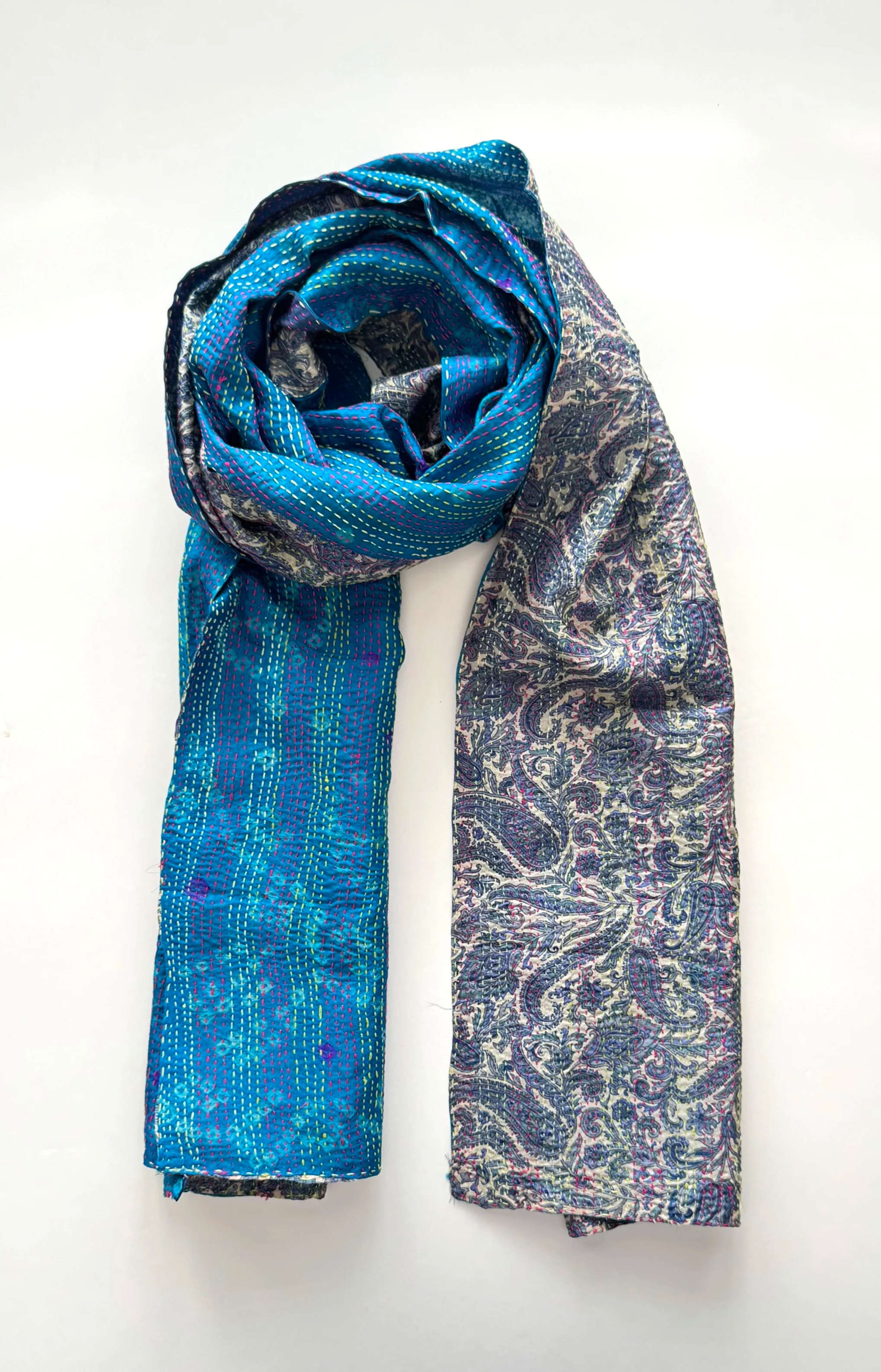 A silk scarf with intricate blue patterns and floral designs is elegantly rolled on one end, creating a textured, sophisticated look on a white background.