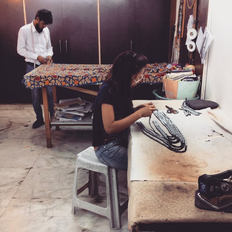 Artisans at work in a studio. A woman sits at a workbench handcrafting necklaces from upcycled sari silk, while a man in the background measures fabric on a patterned table.