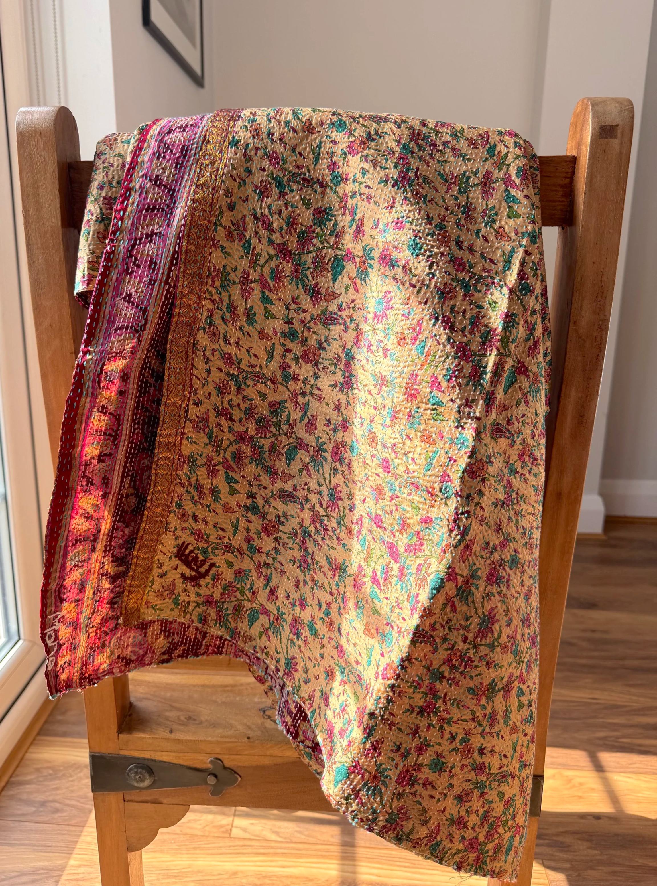 A colourful, intricately patterned quilted silk scarf with floral designs drapes over a wooden chair in sunlight. The vibrant fabric contrasts with the warm wood tones.