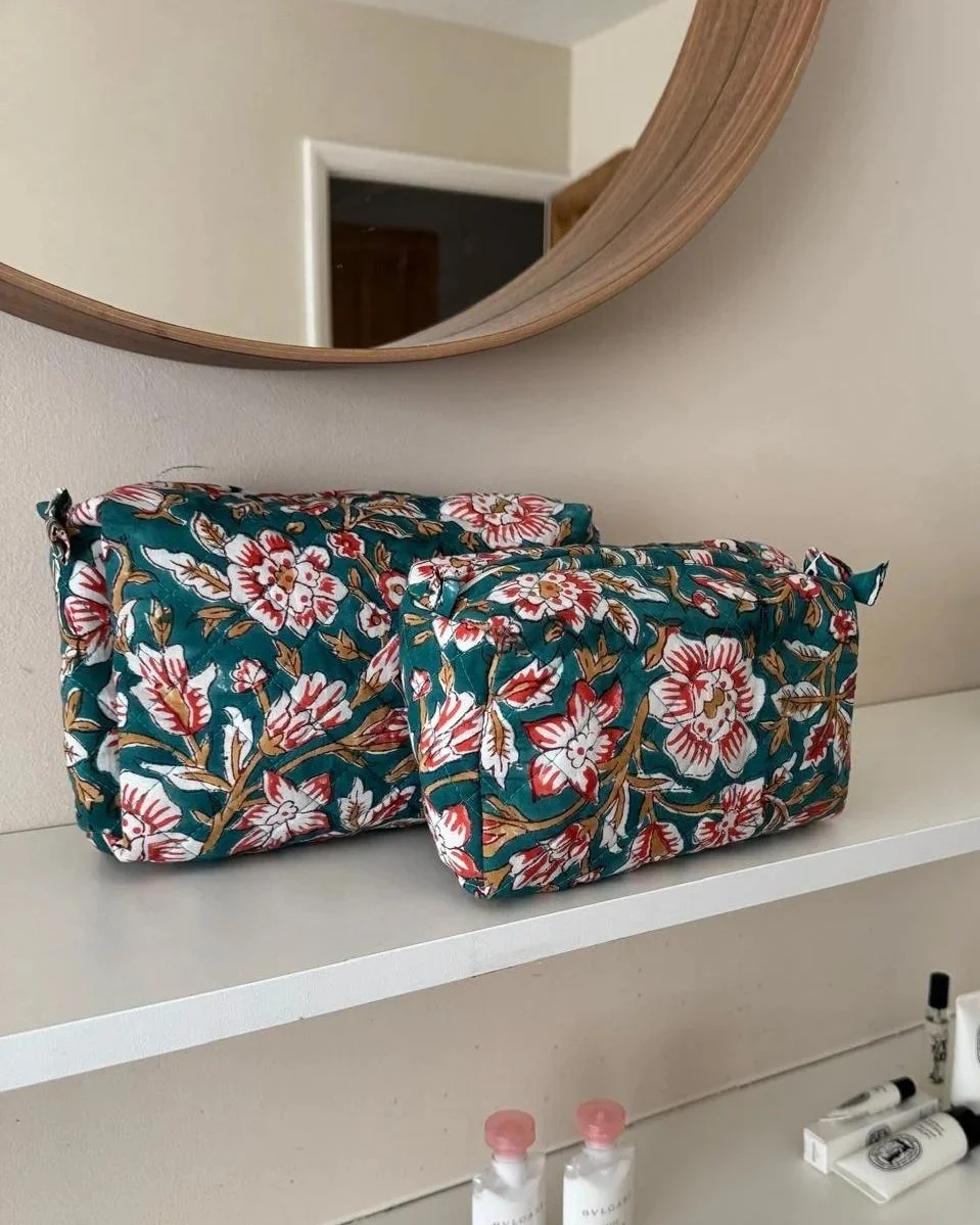 Two block print quilted cotton wash bags resting on a white shelf under a mirror , the wash bags are teal with large white flowers and red highlights