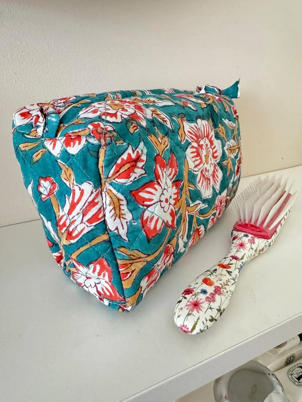 Large teal toiletry travel pouch with red and white flowers resting on a bedroom shelf with a hairbrush resting in front, the wash bag is hand block printed by artisans in India