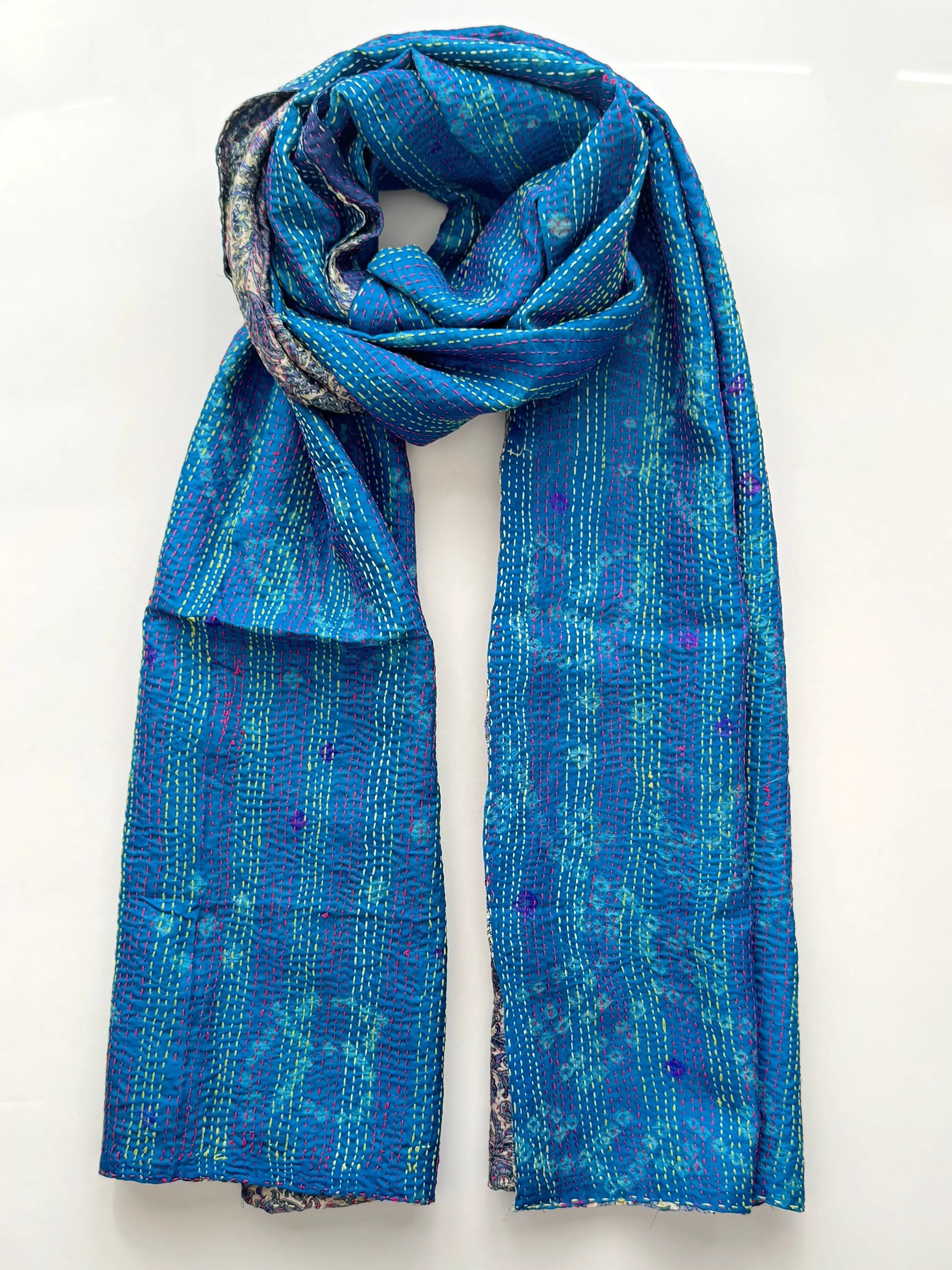 A vibrant handmade blue scarf artfully draped, showcasing intricate Kantha stitching. Its texture and elegance suggest it's crafted from upcycled sari silk and hand embroidered with running stitch.