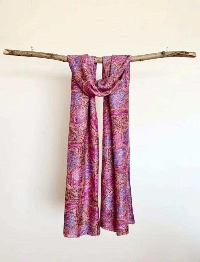 A long pink and purple silk scarf hangs from a wooden branch suspended from a ceiling. The scarf has an abstract patchwork pattern and is embroidered with Kantha stitch to give a quilted texture.