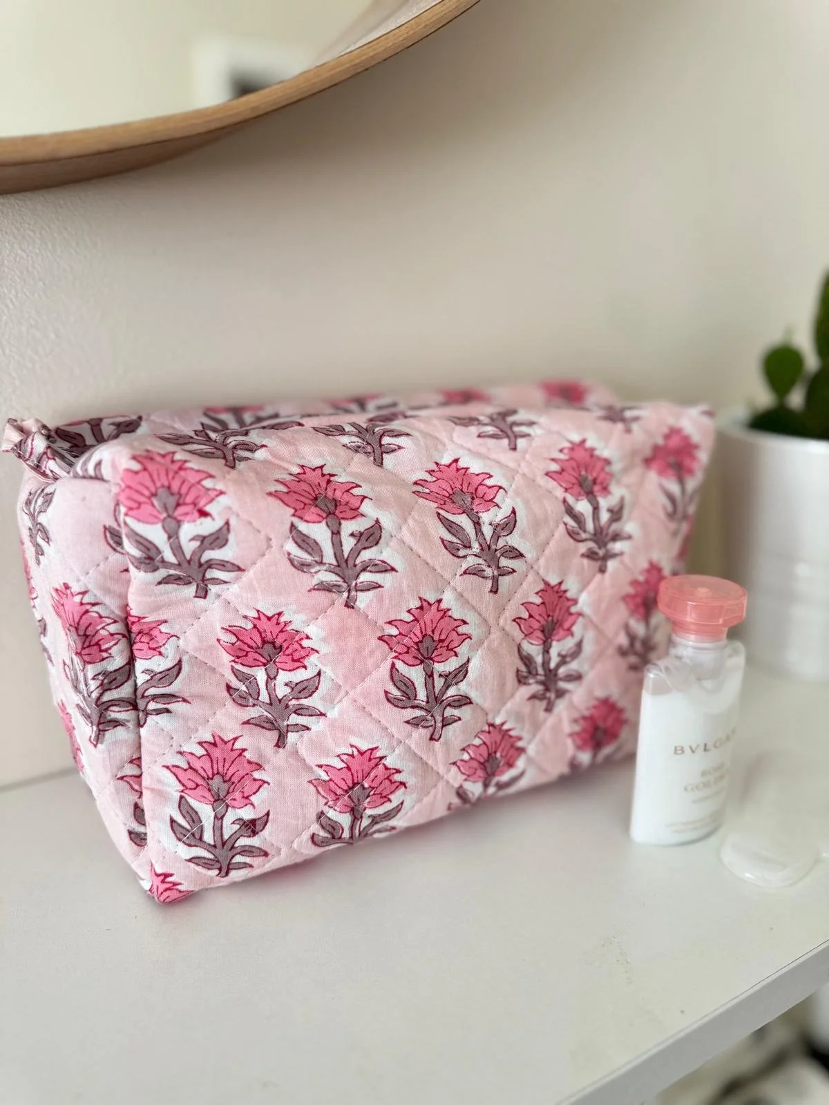 A large quilted cotton pink floral wash bag for ladies resting on a white shelf with a cactus plant in the background.