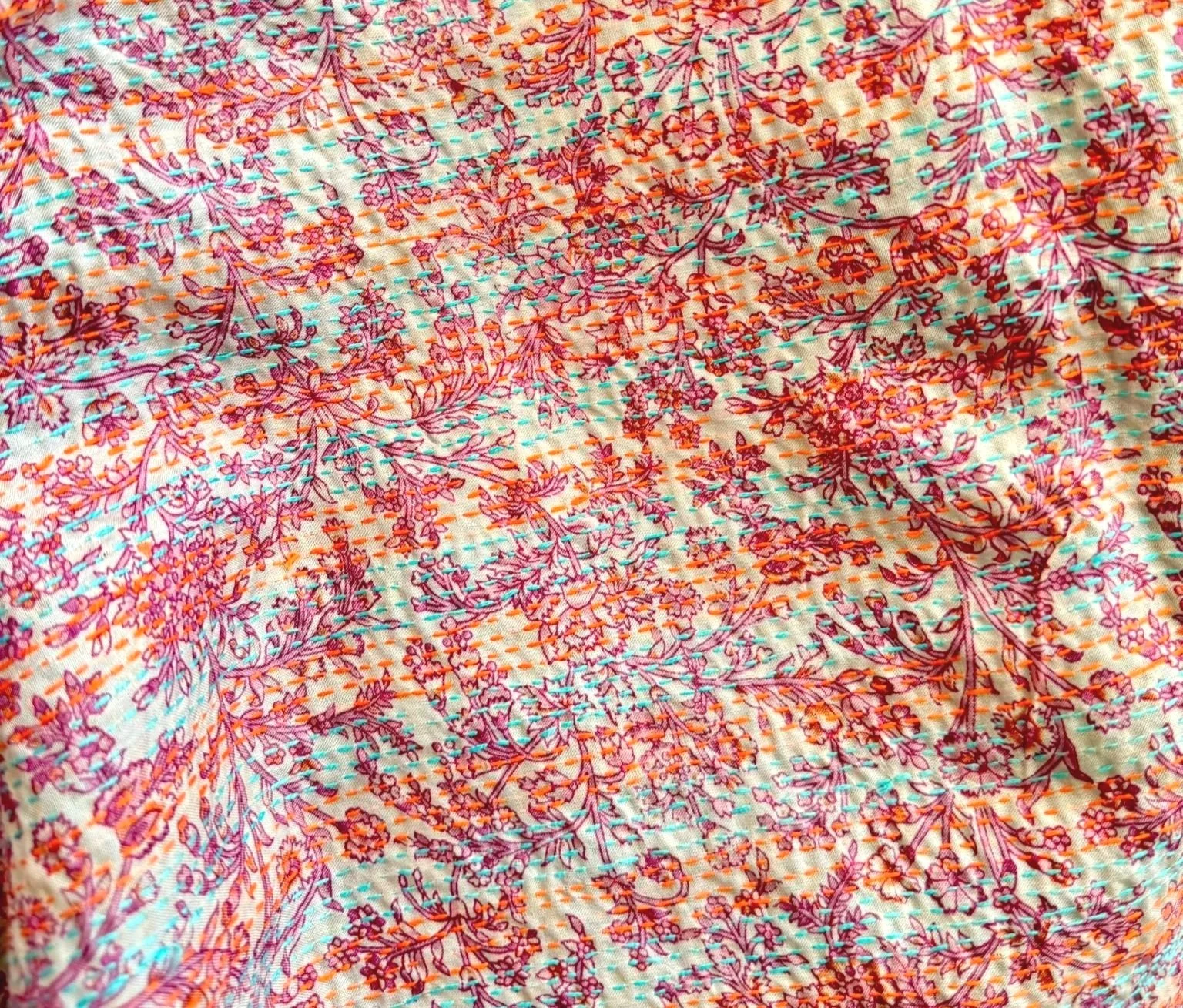 Close-up of a floral textile with a white base, featuring dense, all-over patterns of purple and orange flowers with blue-green stitching.