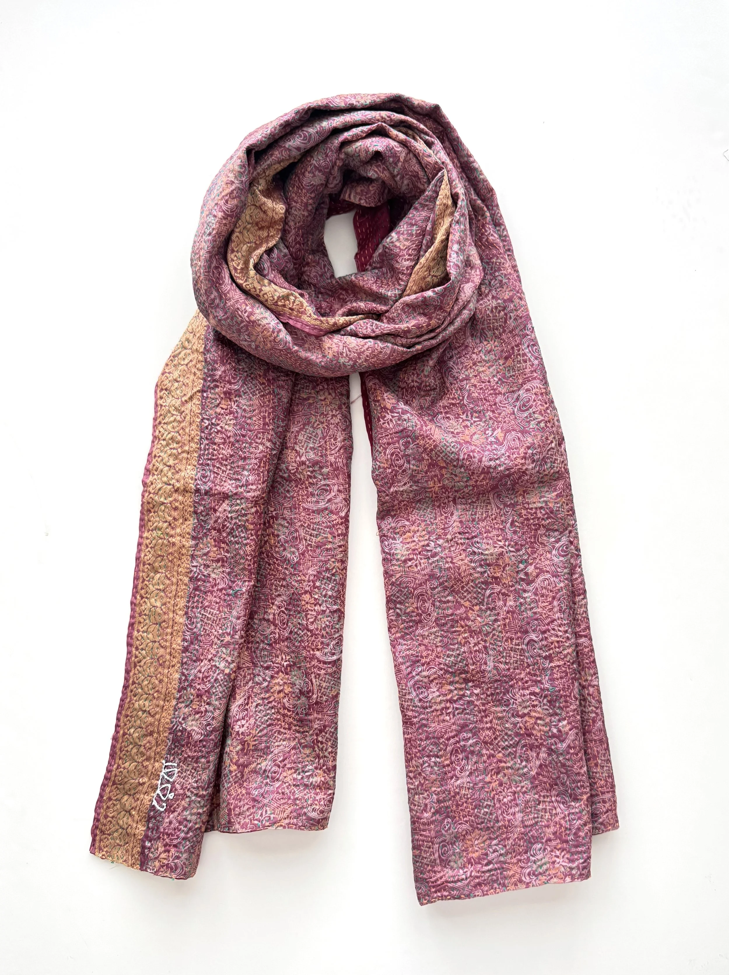 A textured, long scarf with pink and gold patterns is laid out on a white background. The scarf has a soft, cozy appearance with a rustic feel.