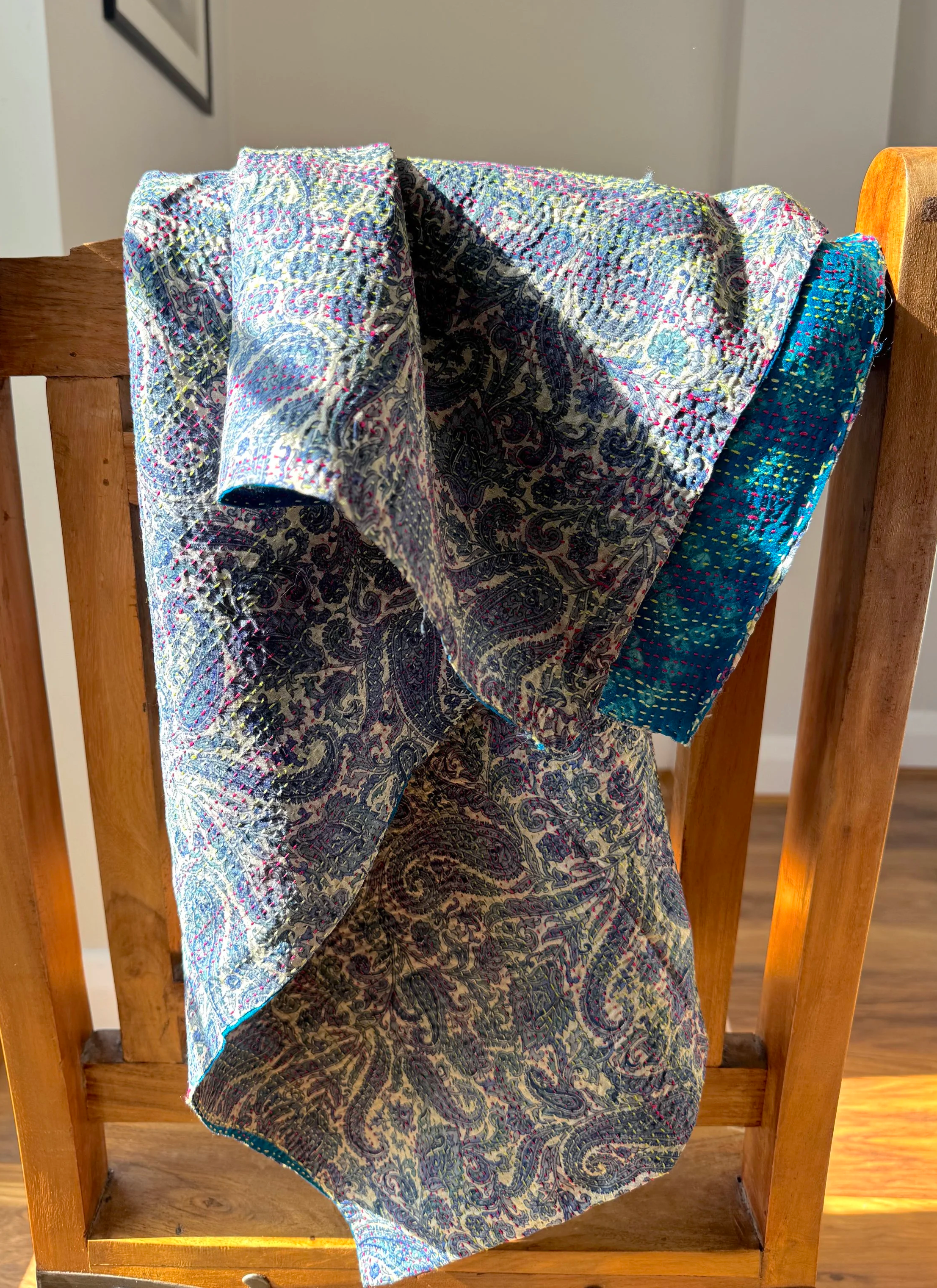 A paisley-patterned kantha sari scarf draped over a wooden chair, bathed in warm sunlight. Textured fabric suggest it is a Kantha sari  hand embroidered scarf.