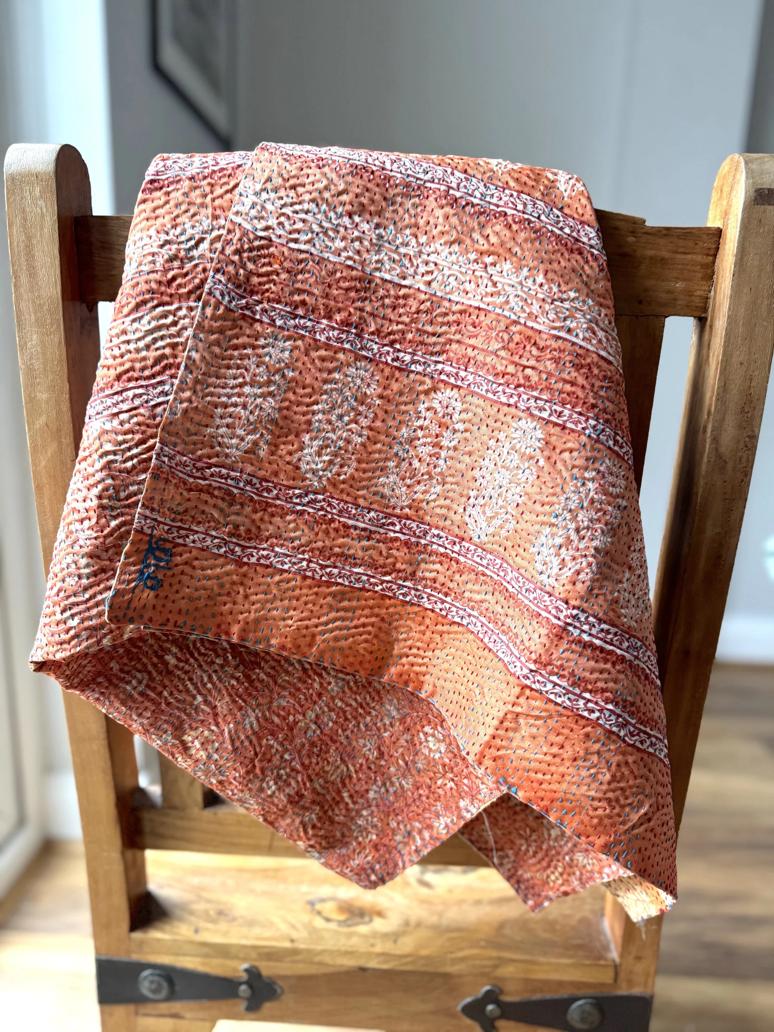 A colourful peach kantha sari scarf draped over a wooden Jali style chair, the scarf has the name of the artisan embroidered in red and features hand embroidered kantha stitch all over