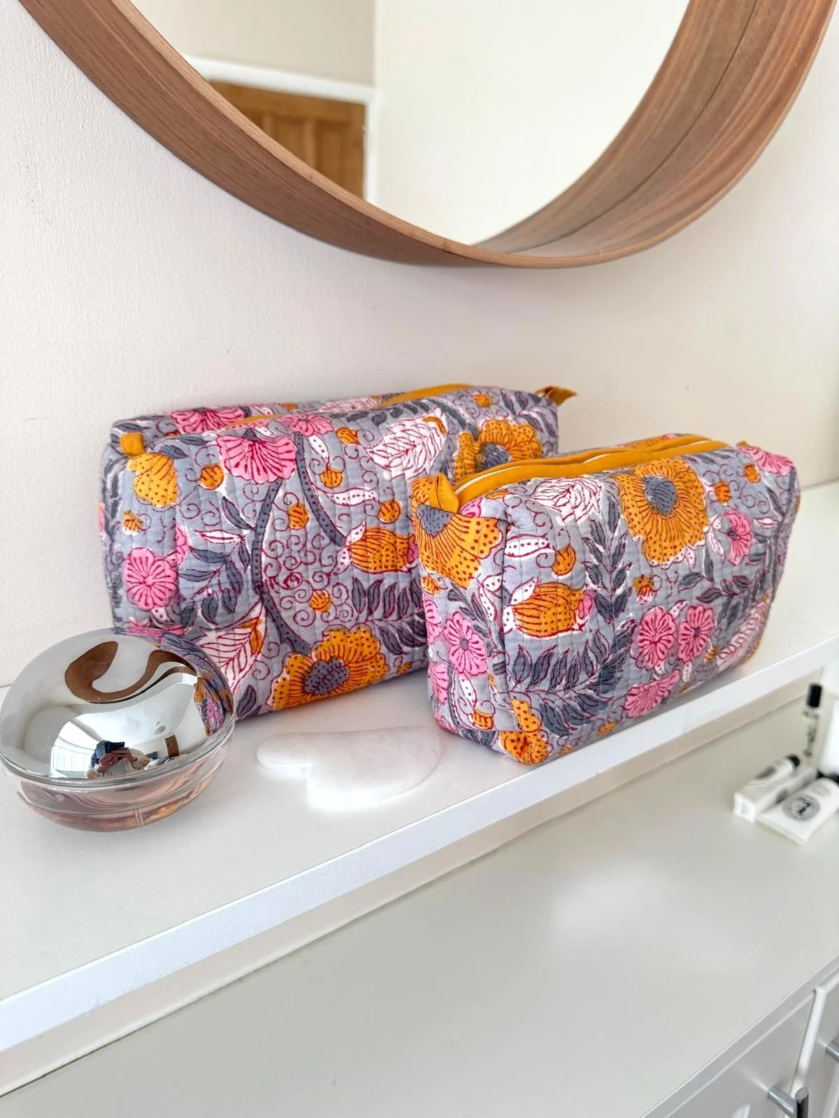 Two handmade block print wash bags in grey, pink and yellow floral print, quilted cotton, sitting on a white dressing table with mirror - Indian artisan cosmetic bags by Jewelled Buddha