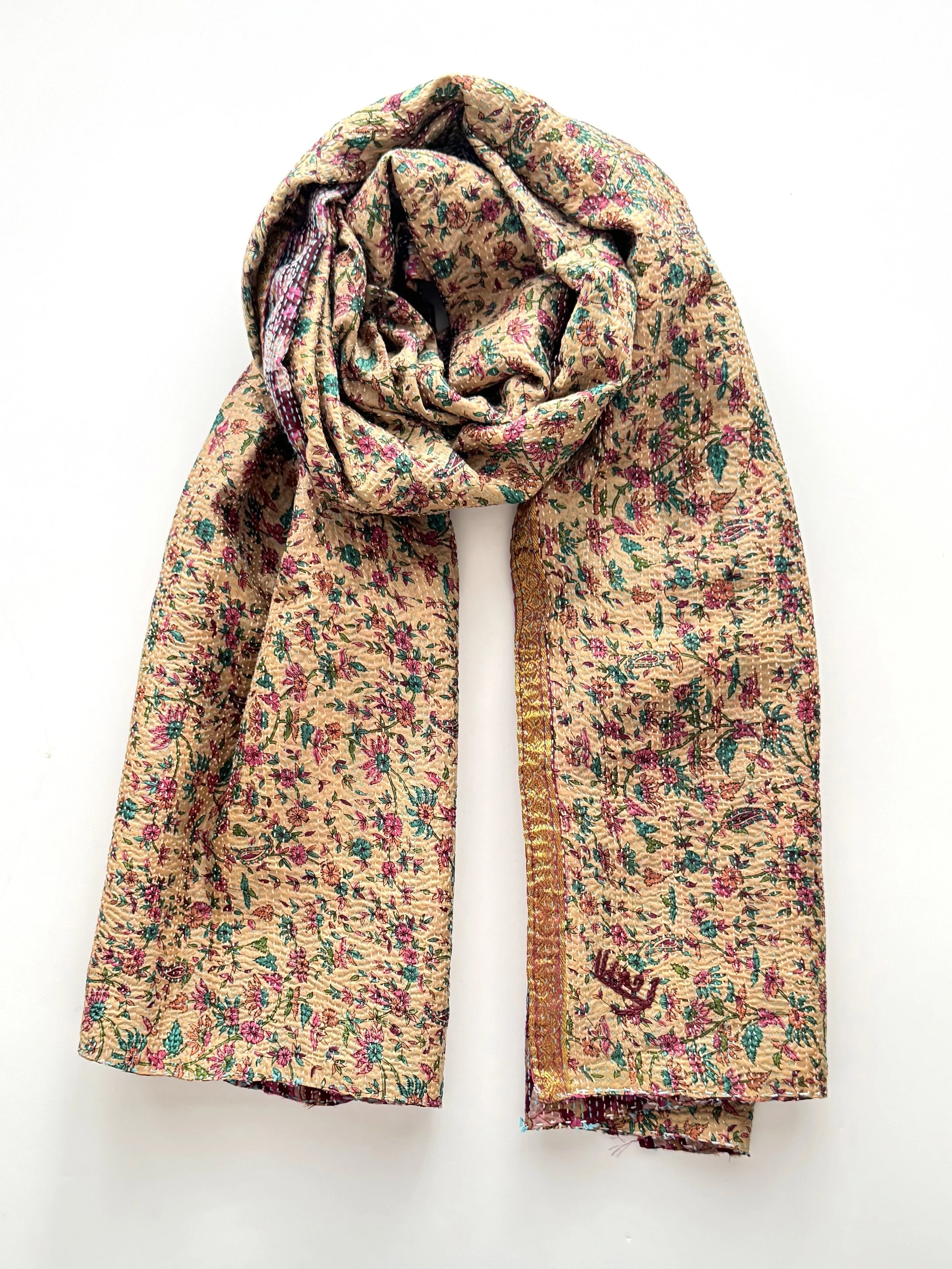 A colorful floral-patterned scarf with pink and green accents on a beige background, elegantly draped against a plain white backdrop.