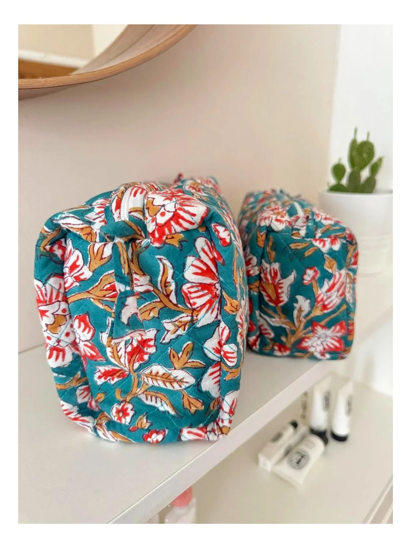 Close up of teal make up bags with large white and red flowers resting on a white shelf with toiletries blurred in the background