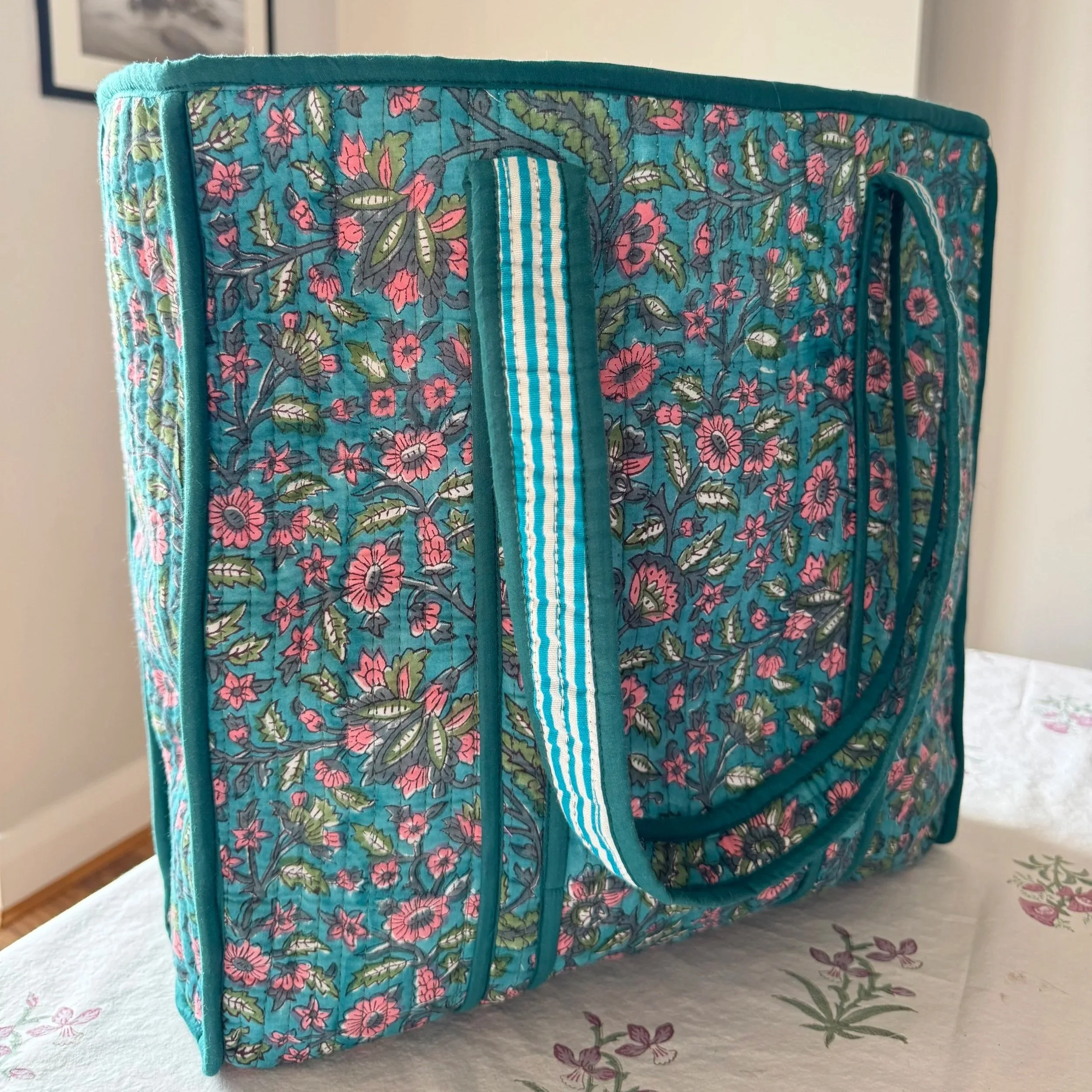 A colourful blue handmade block printed quilted cotton tote bag with colourful pink and green flowers, contrasting blue and white striped shoulder straps and teal green trims.