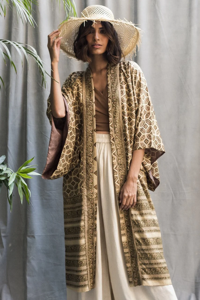 Model wearing a silk kimono robe in beige with a straw beach hat against a beige background