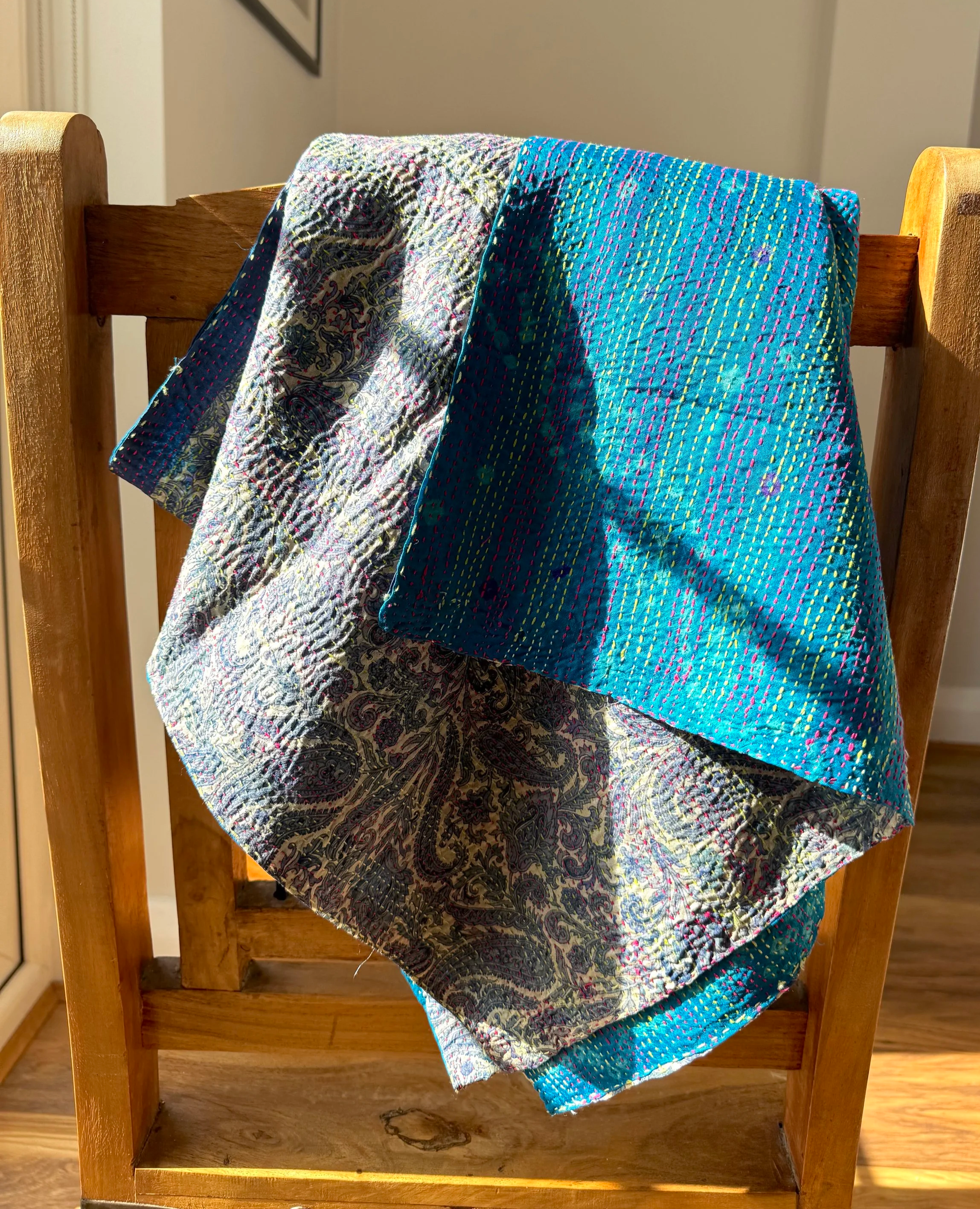 A patterned blue silk scarf with a reversible paisley pattern draped over the back of a wooden chair, bathed in warm sunlight.