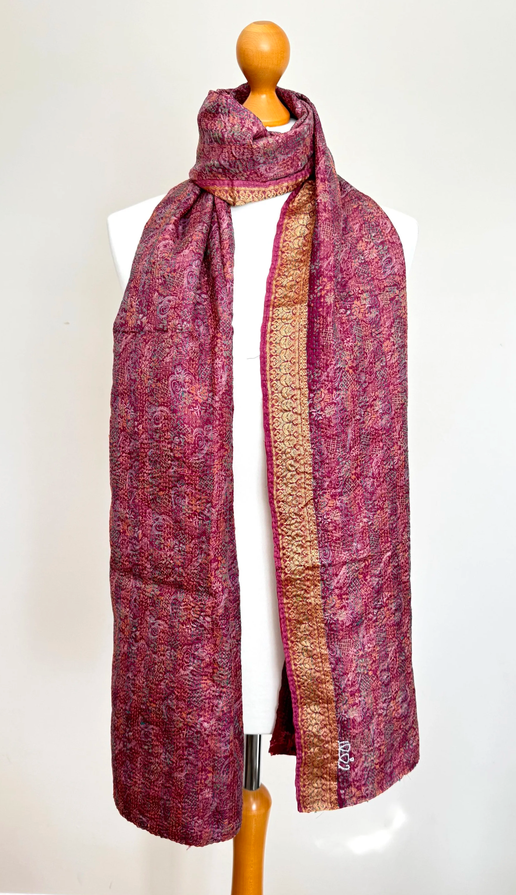A mannequin displays an ornate red and gold shawl with intricate patterns, neatly draped over its shoulders. The background is a plain light wall.