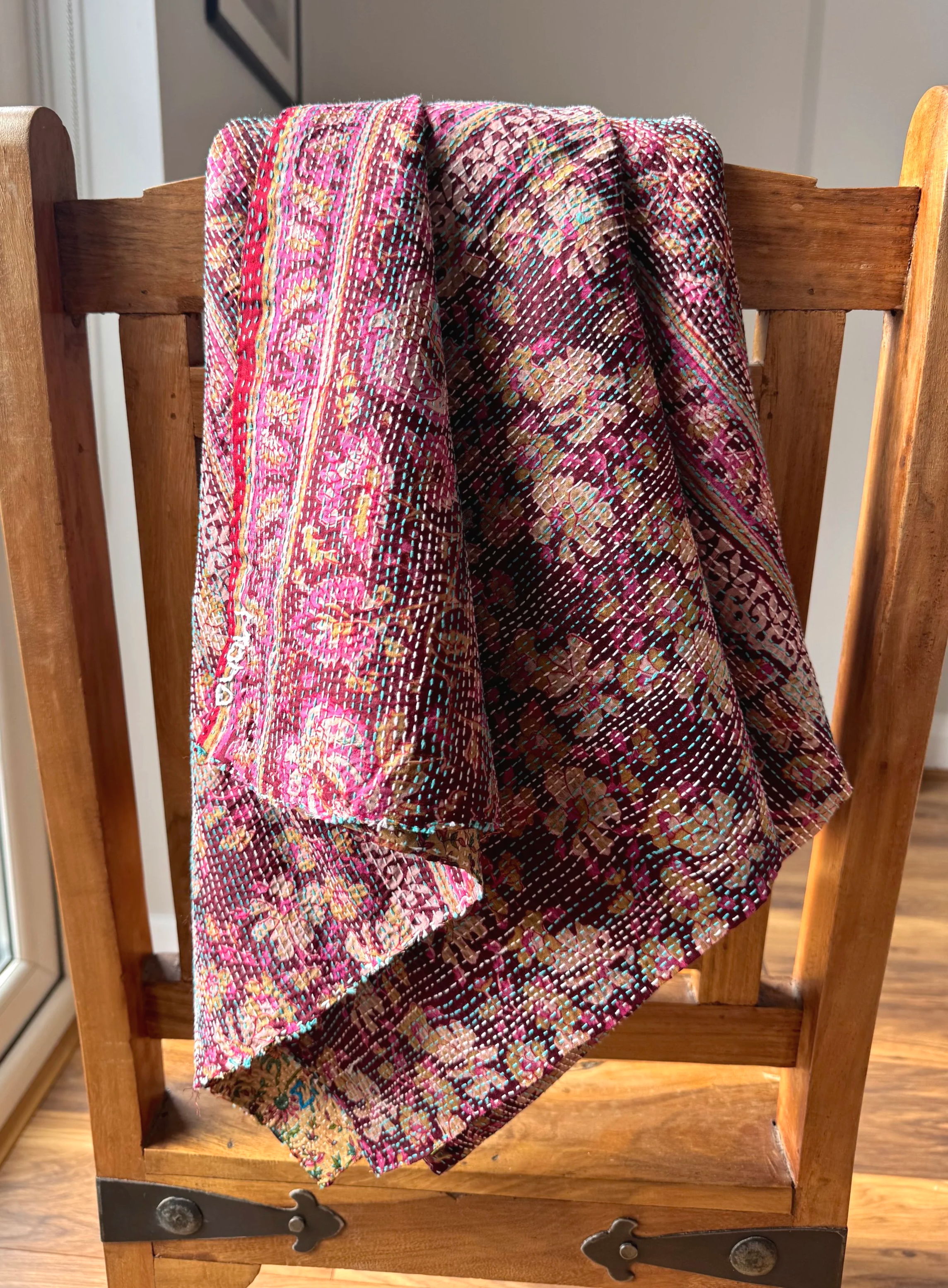 A floral-patterned, colorful quilt hangs over an antique wooden chair by a window, with sunlight streaming in, creating a warm, cozy ambiance.