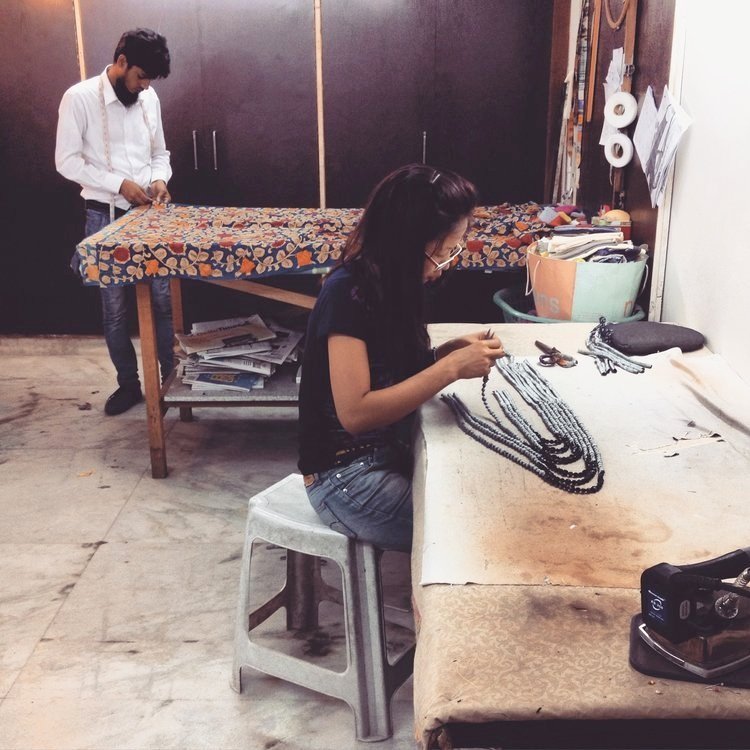 Artisans at work in a studio in India. A woman sits at a workbench handcrafting upcycled sari silk necklaces , while a man in the background measures fabric on a patterned table.